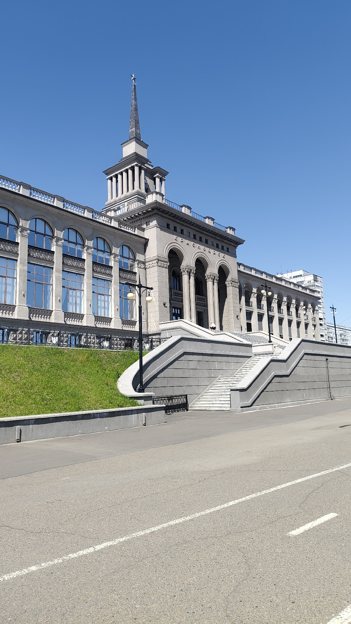 The image displays a grand, neoclassical-style building under a clear, bright blue sky in Krasnoyarsk, Russia. The building's facade features numerous columns, large arched windows, and ornate railings, suggesting a significant public structure, possibly a station or administrative building. A prominent central tower culminates in a slender spire topped with a star, characteristic of Soviet-era architecture.
A large, grey stone staircase with white steps leads up to the building's entrance. To the left of the staircase, a vibrant green grassy slope adds a natural touch to the urban setting. A black street lamp with two lights stands near the base of the stairs. In the immediate foreground, a paved road with a white painted line indicates a street or highway. Another multi-story, more modern building is visible in the background to the right.
The scene is captured during bright daylight, with strong sunlight creating clear shadows, suggesting it is midday or early afternoon. No people or vehicles are visible, leaving the focus entirely on the architecture and landscape. The visible Cyrillic text "КРАСНОЯРСК" (Krasnoyarsk) is prominently displayed on the building's facade, confirming its location. The image conveys a sense of grandeur and historical significance within an urban environment.