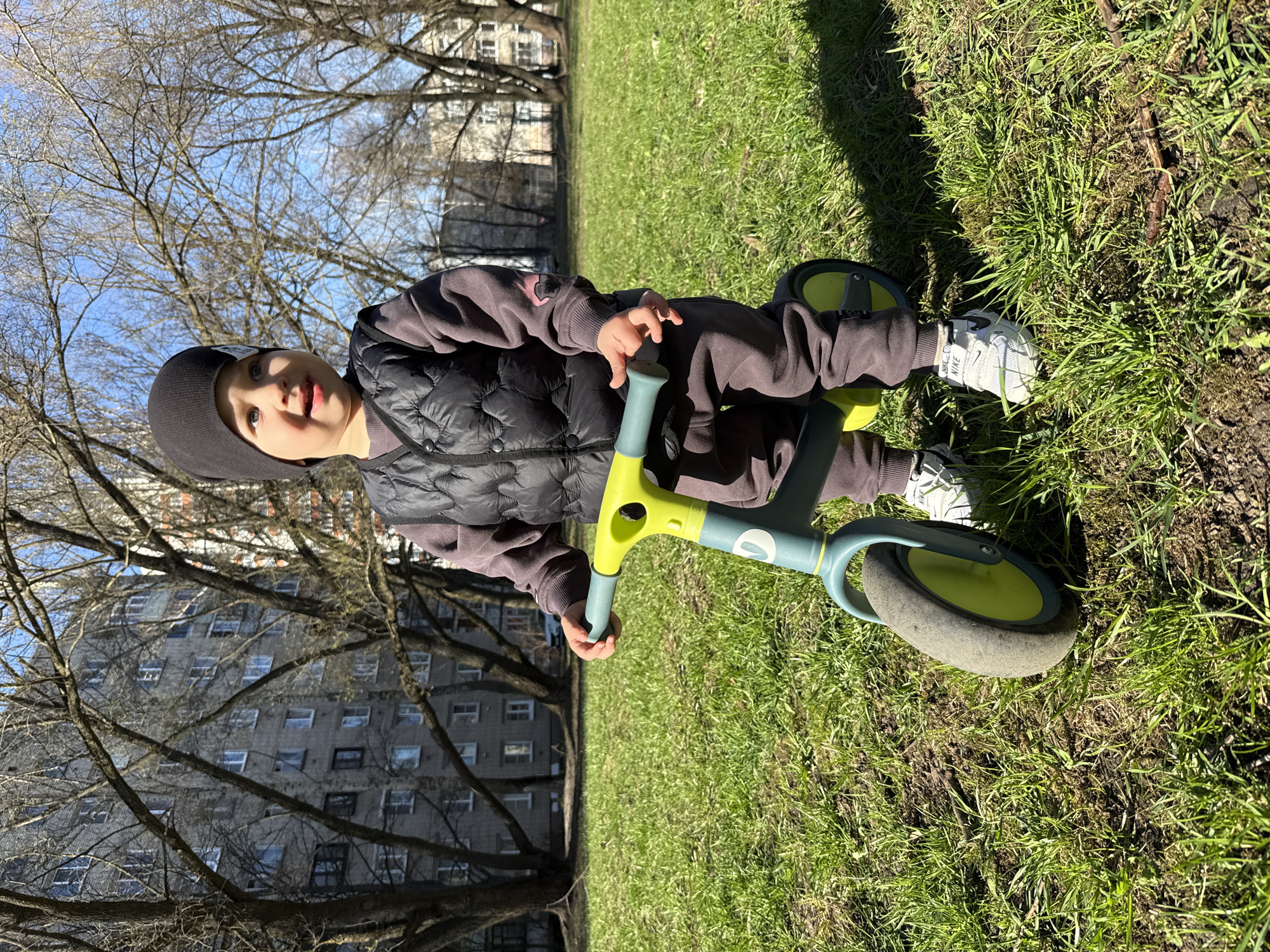 A small child is sitting on a light-blue and yellow tricycle on a grassy lawn. The child is wearing a dark-colored hat, a brown sweater, a black quilted vest, and white sneakers with a light-grey Nike logo. In the background, there are trees without leaves and a grey multi-story apartment building. The location is possibly Rusanovo, Russia.