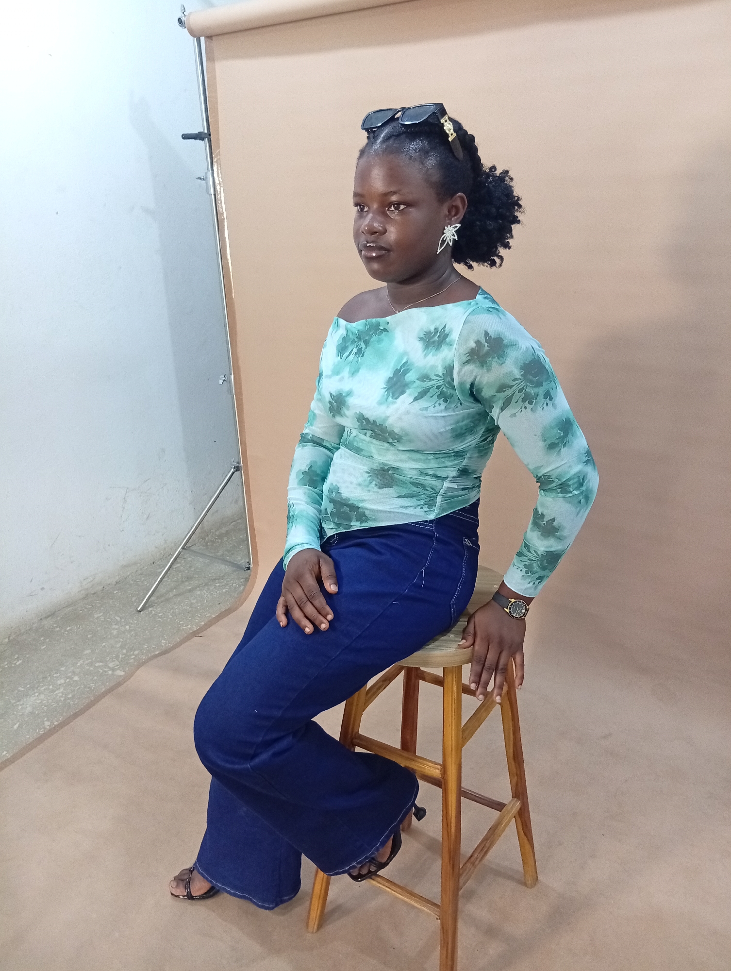 A young woman is seated on a wooden stool in what appears to be a studio. She is looking to her left and has sunglasses perched on top of her head. She is wearing a light green, off-the-shoulder, long-sleeved top with a floral pattern and dark blue flared jeans. A wristwatch is visible on her left wrist. The background consists of a beige backdrop. To the left of the backdrop, a white wall with a lighting stand is visible. The floor is a neutral color. The image does not provide any specific information that would allow for a precise location within the city of Gura Top, Nigeria.