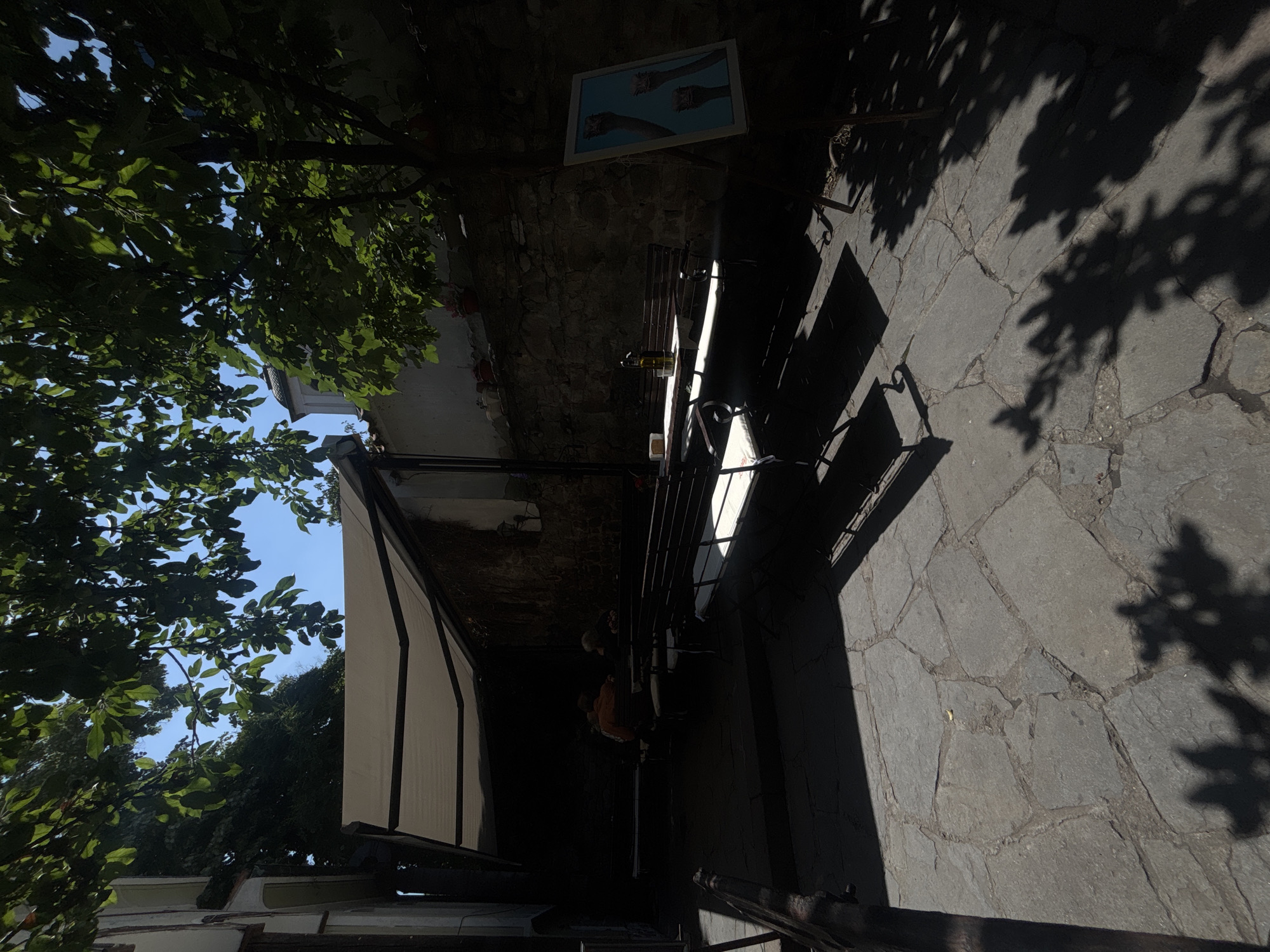 The image depicts an outdoor seating area, likely a cafe or restaurant patio, on a bright and sunny day in Plovdiv, Bulgaria. The perspective is slightly elevated, looking down into the space.
On the left side of the frame, vibrant green tree foliage is prominent, with sunlight filtering through the leaves and revealing patches of clear blue sky. The sunlight casts dynamic, dappled shadows of leaves and branches onto the stone-paved ground below and the adjacent structures. The ground itself is made of irregular grey stone slabs.
In the mid-ground, a dark stone wall serves as a backdrop. A white-framed picture hangs on this wall, showcasing an image of two hands holding what appear to be ice cream cones or similar desserts against a light blue background. Below the picture, a dark outdoor table or bench is positioned, with parts of its surface brightly illuminated by sunlight. On the table, objects like bottles and perhaps glasses or plates are visible in the sunlit patches. Two individuals are seated at this table, though their details are largely obscured by shadows, with one person wearing an orange top.
To the right, a large white and black striped awning or canopy extends over a section of the outdoor area, providing shade. The strong contrast between bright sunlit areas and deep shadows suggests a clear, sunny day, likely during the morning or afternoon. The overall scene evokes a sense of calm and leisure, typical of enjoying an outdoor meal or drink in a charming, historic urban setting. No text is explicitly visible other than the content of the framed picture.