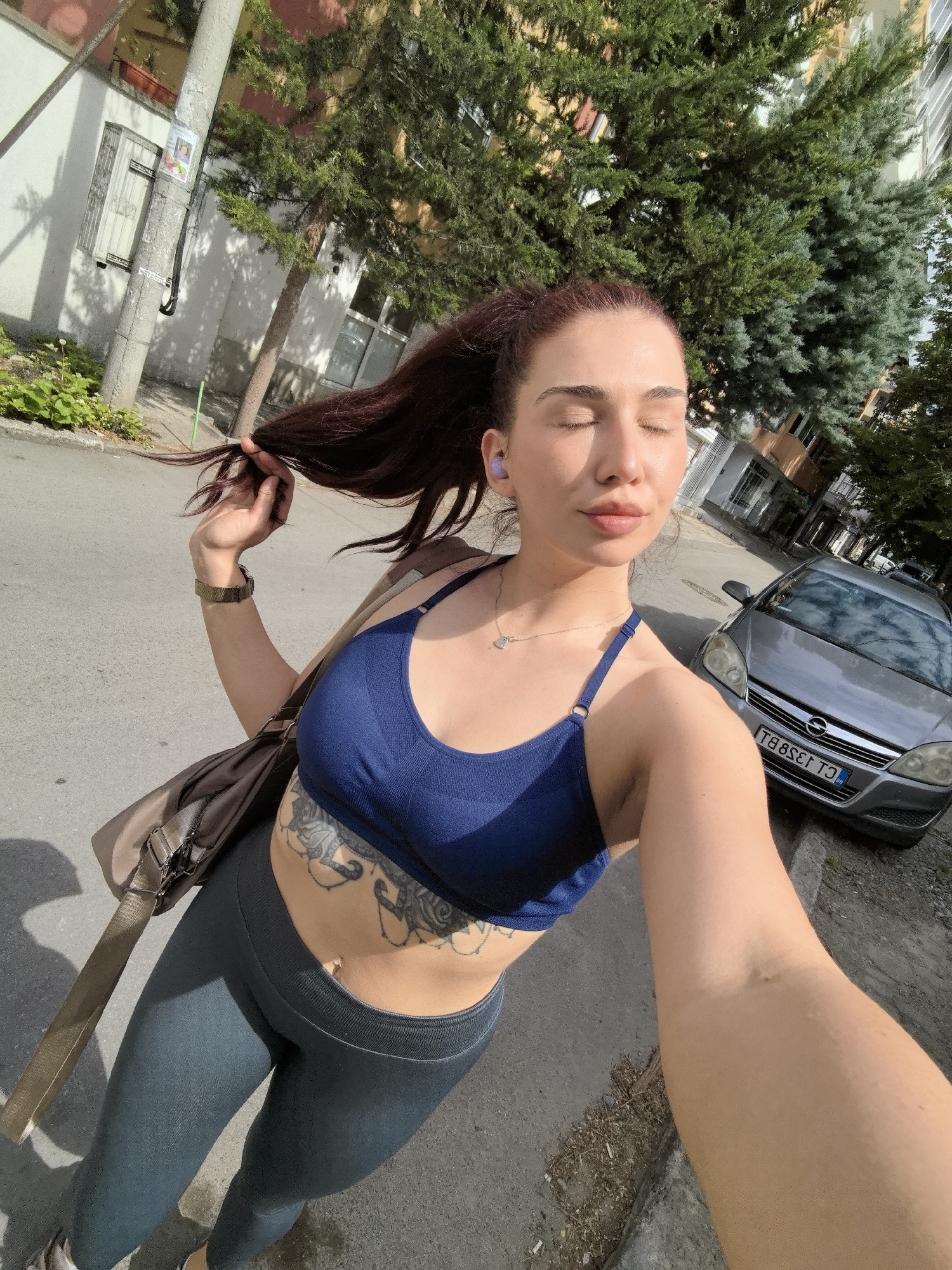 A woman stands on a street in Stara Zagora, Bulgaria, holding a strand of her long, reddish-brown hair. She is wearing a blue sports bra and gray leggings. A tattoo is visible on her abdomen. She has a shoulder bag and a watch on her left wrist. Her eyes are closed, and she has wireless earbuds in her ears. A gray Opel Astra car is parked on the side of the road. The street appears to be residential, with buildings and trees in the background. The license plate of the Opel Astra car is "TB 8581 CT."