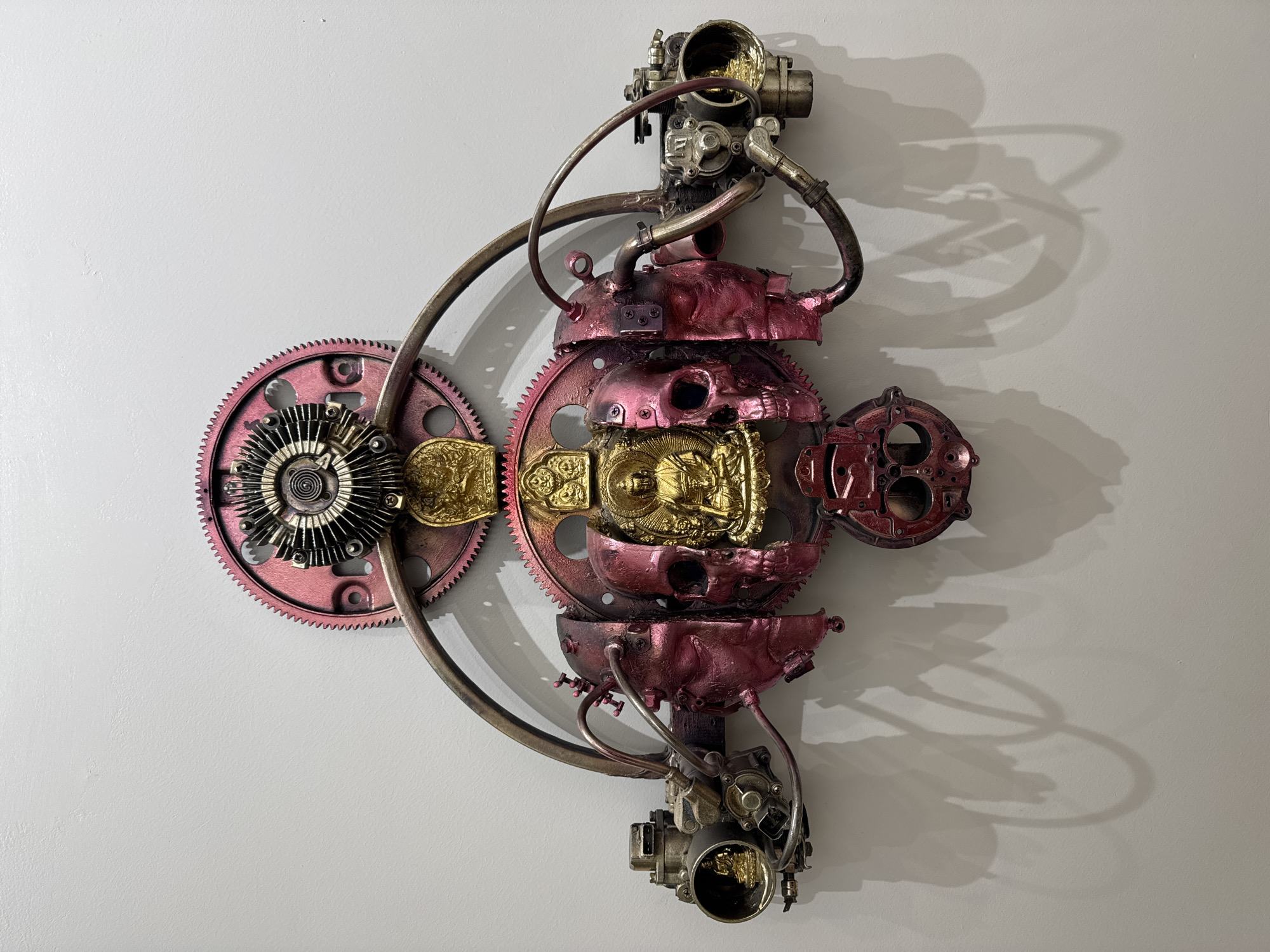 A metal sculpture hangs on a white wall. The sculpture is constructed from various mechanical parts, including gears, pipes, and engine components. The parts are painted in shades of pink, gold, and silver. The sculpture has a roughly symmetrical shape and is oriented vertically. A shadow of the sculpture is cast on the wall to the right. The environment is a plain white wall, suggesting an interior space. The location in Ulaanbaatar, Mongolia cannot be determined from the image alone.
