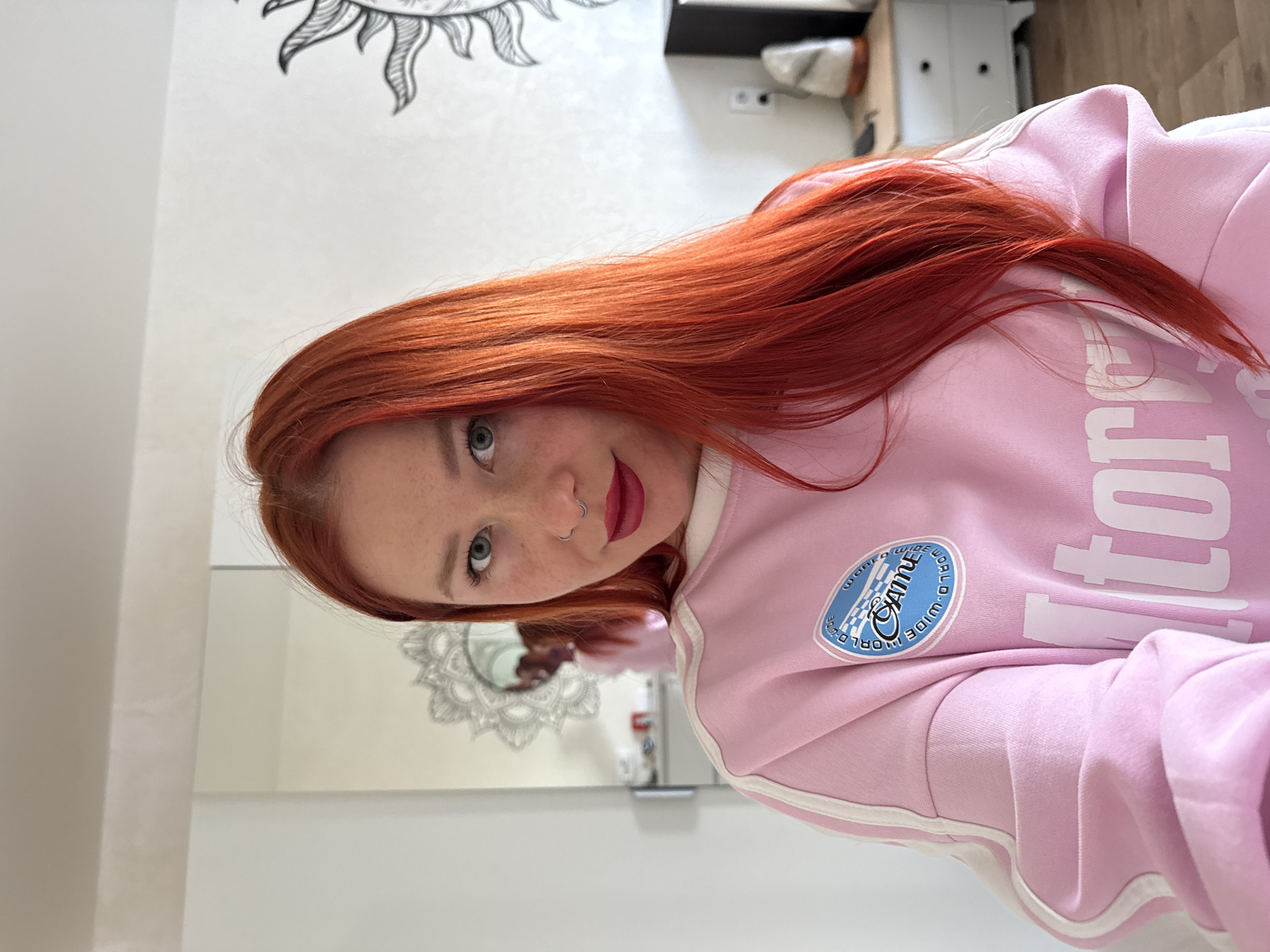 A young woman is featured in a close-up, rotated portrait, her head positioned towards the left of the frame. She possesses long, vibrant red hair that flows over her shoulders and part of her attire. Her light blue-green eyes are directed straight at the viewer, and her face is adorned with freckles across her nose and cheeks. She wears a silver hoop nose ring in her left nostril and bold red lipstick. Her expression appears calm and direct.
She is dressed in a light pink hooded sweatshirt featuring white piping along the seams of the sleeves and waist. A circular patch on her left chest displays a blue and white checkered pattern, with the English phrase "world wide" and the Russian word "CAMOE" (meaning "the most") in white stylized text. Partial white lettering, possibly forming "tory" or "itory" as part of a larger word like "Victory," is also visible on her sweatshirt.
The setting is an indoor environment, likely a contemporary room in a residence in Rostov-on-Don, Russia. The background consists of white walls. A black and white stylized sun motif is partially visible on the upper left wall. Below her, a large mirror reflects an ornate, mandala-like decorative element. Other background details include a white cabinet, a dark shelf, and an electrical outlet, indicating a domestic scene. The lighting is bright and even, suggesting daytime. The image has been rotated 90 degrees clockwise from its original orientation.