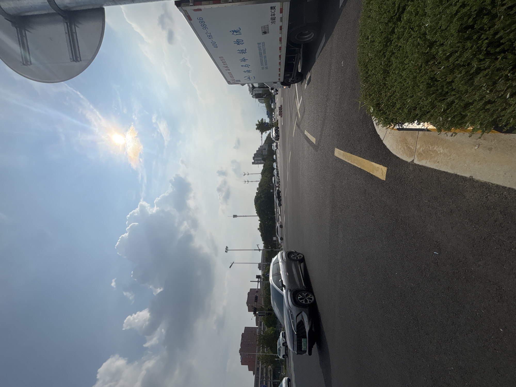 The image depicts a sunlit urban intersection in Suzhou Industrial Park, China. The sky is a bright blue with scattered white clouds, and the sun is partially obscured by a cloud, creating a lens flare effect. A large, round traffic signpost is visible in the upper right corner. In the foreground, a grey car with a license plate reading "豫883525" is turning left at the intersection. A white delivery truck with Chinese text and a phone number "400-182-9886" is parked on the right side of the road. The road surface is asphalt with yellow lane markings. In the background, several buildings, including multi-story brick structures, are visible, along with trees and distant utility towers. The overall scene suggests a busy, modern urban environment under a clear sky.