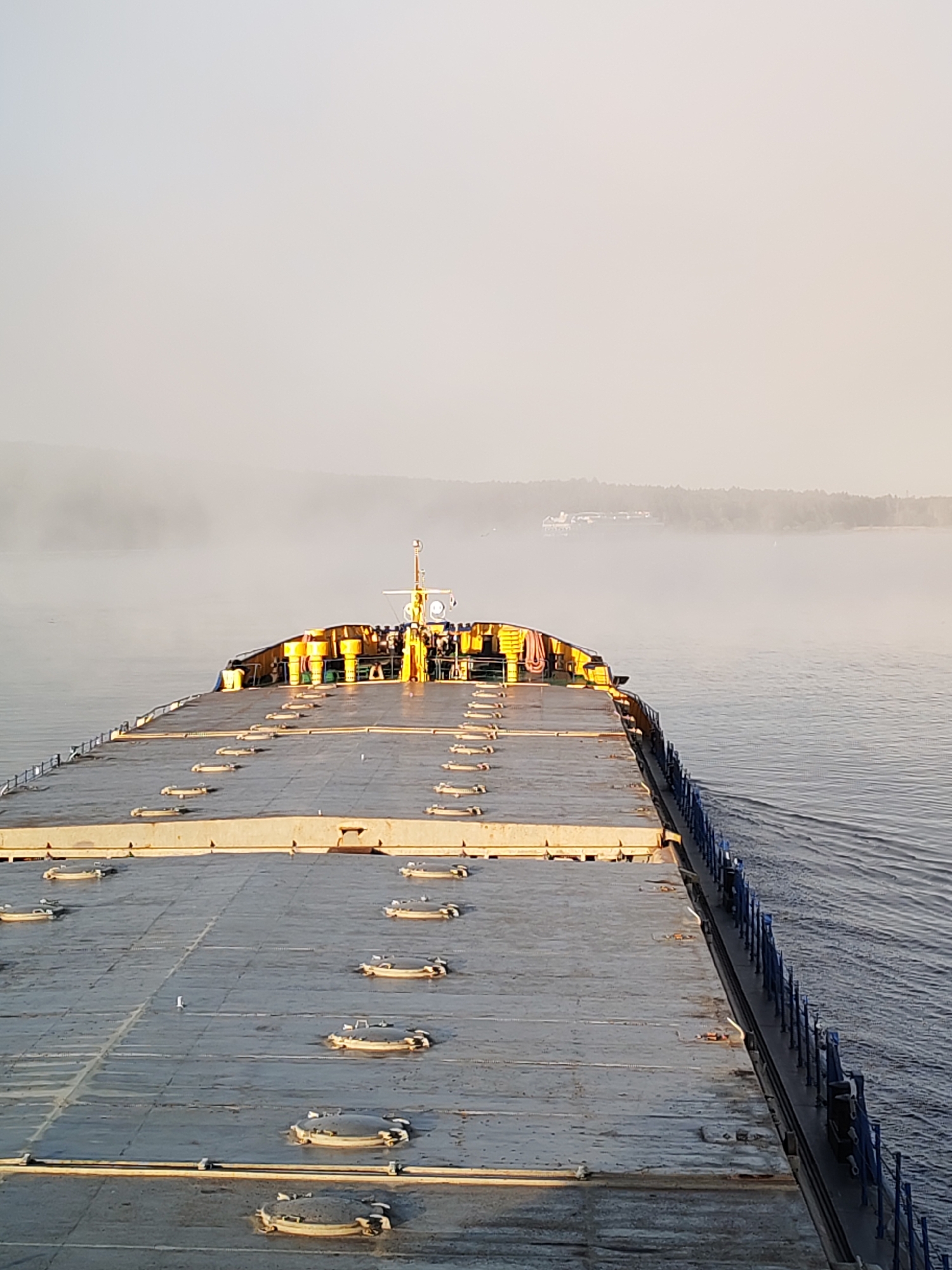 The image displays a large, flat-decked vessel, likely a cargo barge or bulk carrier, observed from a high vantage point looking forward from its stern. The deck, predominantly grey and appearing somewhat damp or worn, is marked by numerous rows of oval-shaped, raised hatches. Towards the vessel's bow, a distinct yellow superstructure or bridge section is visible, featuring a mast equipped with a radar dome and antenna. Several small figures, presumably crew members, are discernible on this yellow structure. Yellow cylindrical objects are arranged along the upper perimeter of this section. A blue railing or fence runs along the port side of the vessel. The vessel is actively moving through calm, dark blue water, generating a wake visible along its sides. The background features distant land covered with trees, significantly obscured by a pervasive atmospheric haze or fog that extends across the water. Faint silhouettes of other vessels are barely visible in the far distance within this haze. The sky is a uniform, desaturated light grey, indicating diffused light conditions. The specific location is Unknown, Unknown.