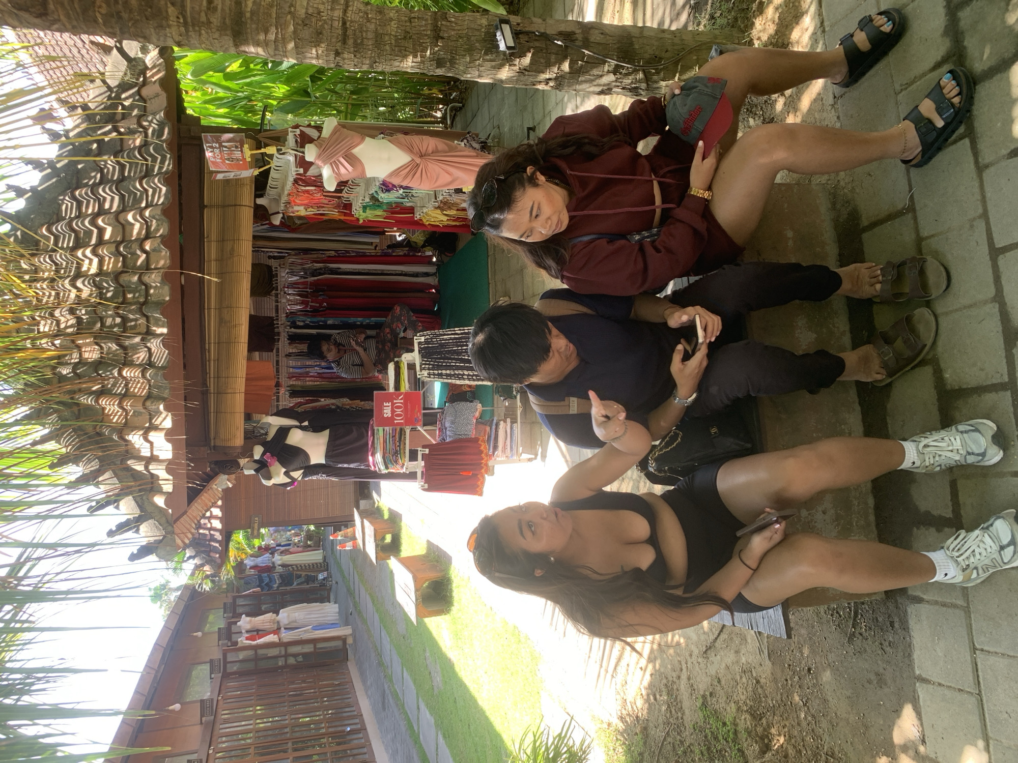 In North Kuta, Indonesia, three individuals, two women and one man, are seated on a wooden bench in front of an outdoor market stall. The woman on the left wears a black bikini top and shorts, her left hand gesturing towards the man. The man in the center is wearing a dark blue t-shirt and black pants, looking down at his phone. The woman on the right is in a maroon hoodie and shorts, holding a baseball cap. The stall behind them displays various clothing items, including swimsuits and dresses, with a "SALE 100K" sign visible. Palm trees and lush greenery surround the scene, with paved ground underfoot. The lighting suggests a bright, sunny day.