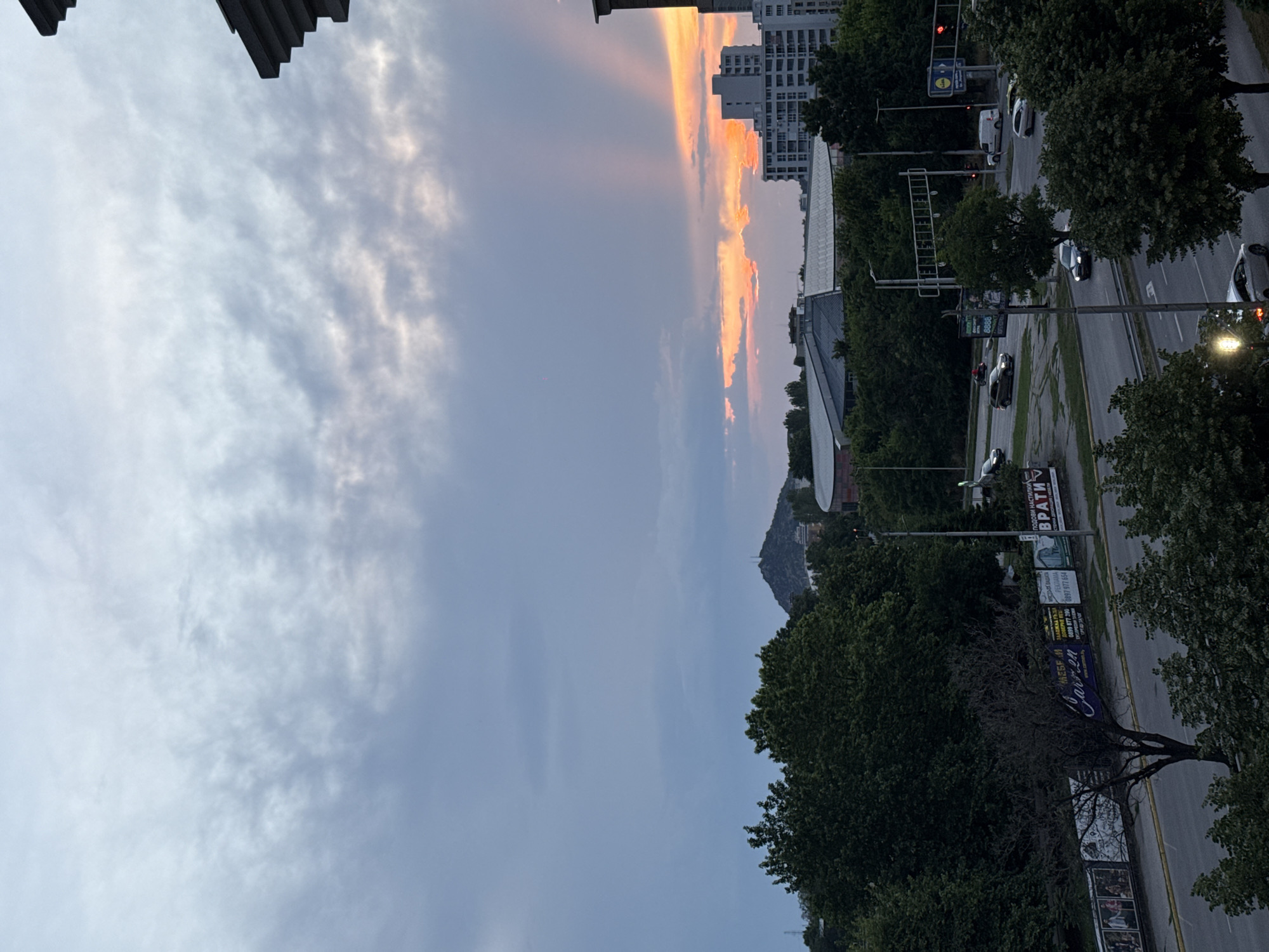 This image captures an urban landscape during sunset or late dusk in Plovdiv, Bulgaria. The sky is a prominent feature, displaying a striking contrast: the left side is filled with soft, textured grey and white clouds, while the right side glows with intense orange, pink, and yellow hues, indicative of the setting sun, with visible crepuscular rays.
The scene includes a multi-lane road running through the city, with several cars visible, some showing illuminated headlights, suggesting the transition from day to night. Streetlights are also active, casting light along the roadway. On the right, various multi-story buildings with numerous windows rise, and a large building with a distinctive, curved roofline (possibly a stadium or arena) is visible in the mid-ground.
Lush green trees line the streets and are scattered throughout the scene, providing a natural element. In the distance, a prominent hill or mountain, partially covered in vegetation, stands against the horizon.
Several commercial signs and billboards are visible along the road. Recognizable text includes the "Lidl" logo, "ПОДЛОГИ НАСТИЛКИ ВРАТИ" (Flooring, Coatings, Doors), "СЕРВИЗ 24" (Service 24) or "Car 24", and various phone numbers such as "0899 977 664" and "8888". The image conveys a serene yet active urban environment as evening approaches.