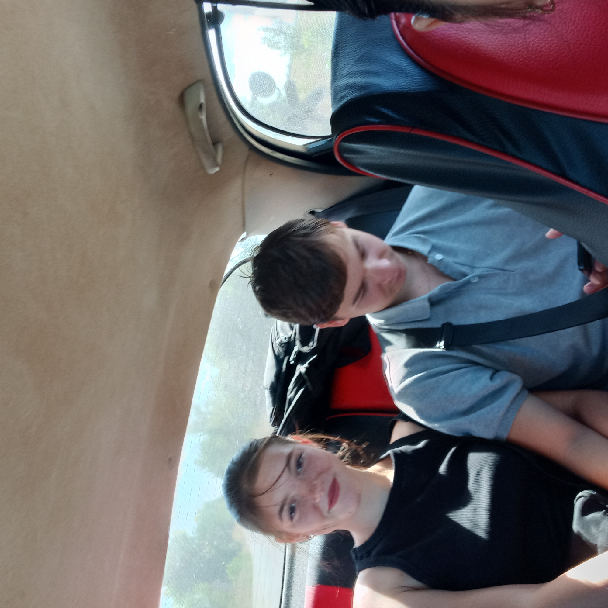 The image depicts two individuals, a young female and a young male, seated in the back of a vehicle, likely a car. The young female, on the left, is wearing a black tank top and has her hair pulled back in a ponytail. She is looking towards the camera with a slight smile. The young male, on the right, is wearing a gray polo shirt and appears to be looking down at a mobile phone in his hand. Both individuals are seated on car seats that have red and black upholstery. There is luggage visible behind the young female. The background outside the car windows suggests a rural or semi-rural setting with trees and vegetation, under a bright sky, consistent with an outdoor environment. The image is taken from within the vehicle, looking towards the back seat. The location of Klyntsi, Ukraine, is not ascertainable from the visual information presented in the image.