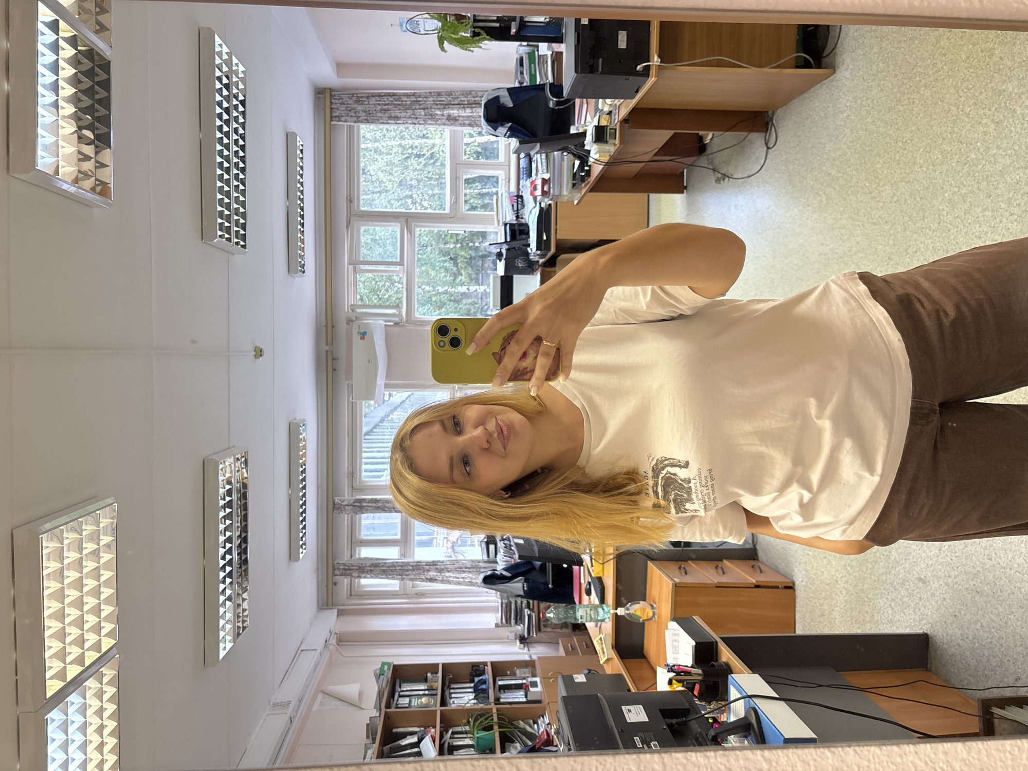 A young woman with blonde hair is taking a selfie in a mirror in what appears to be an office or classroom environment. She is wearing a white t-shirt and brown pants. She is holding a yellow smartphone in her right hand, which is capturing the reflection.
The environment behind her is visible in the mirror. There are desks with computer monitors, office supplies, and shelves filled with files and books. Large windows are visible in the background, with trees and foliage visible outside, suggesting a daytime setting. The ceiling has fluorescent light fixtures. The floor is a light-colored speckled material.
The image is a digital photograph, likely taken with a smartphone. The location is not specifically identified as being in the city of Rusanovo, Russia.