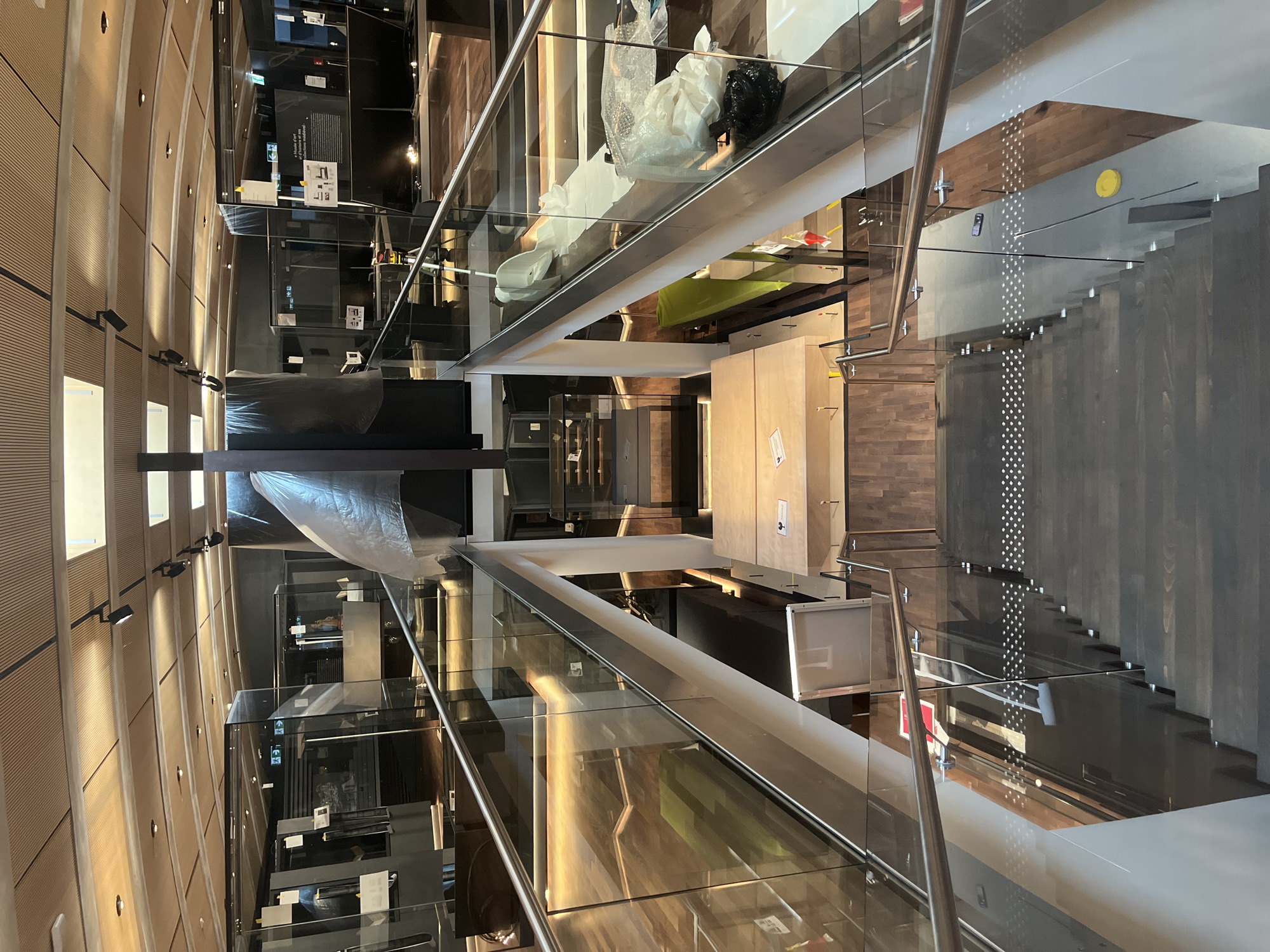 An interior shot looks down a modern wooden staircase with glass railings. The staircase is in a multi-level building, possibly a museum or exhibition space, with the ceiling featuring wooden slats and integrated lighting. Displays behind glass are visible on the upper levels, some covered with protective sheeting. Various objects, including what appear to be large crates, bubble wrap, and black trash bags, are present on the landings and along the sides of the stairwell. The scene suggests a period of setup, renovation, or closure for the space. The image does not provide information to identify the specific city of Cologny, Switzerland.