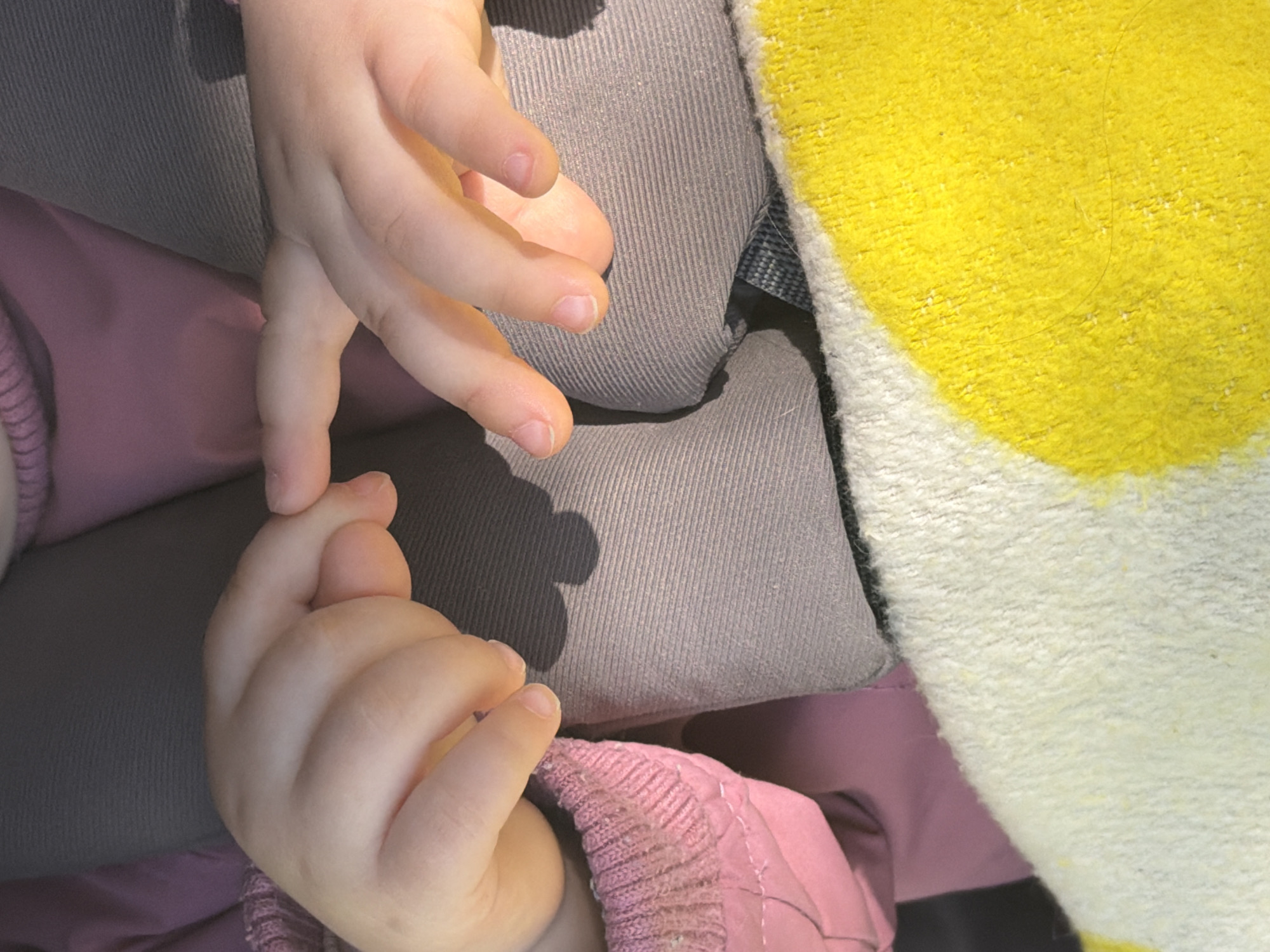 A close-up shot shows a child's hand resting on a gray corduroy surface, partially covered by a yellow and white patterned fabric. The child is wearing a pink jacket. The location, Tokkari, Russia, is not discernible from the visual information provided.