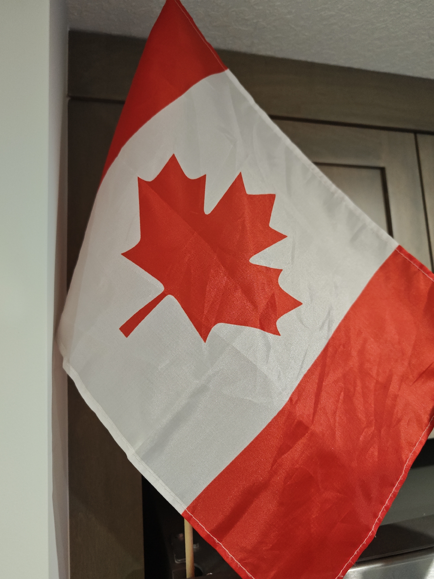 The image shows a close-up of a Canadian flag displayed indoors. The flag is made of fabric and features the iconic red maple leaf on a white background, bordered by red bars on either side. The flag is partially unfurled and hangs at an angle, with some creases and folds visible, suggesting it might be a decorative item or a small handheld flag. The background is blurred but appears to be an interior setting, possibly a room with wooden cabinets or furniture. The lighting suggests it is daytime, but there are no specific time-of-day indicators. The context provided indicates the location is Calgary, Canada. No people or specific events are depicted.