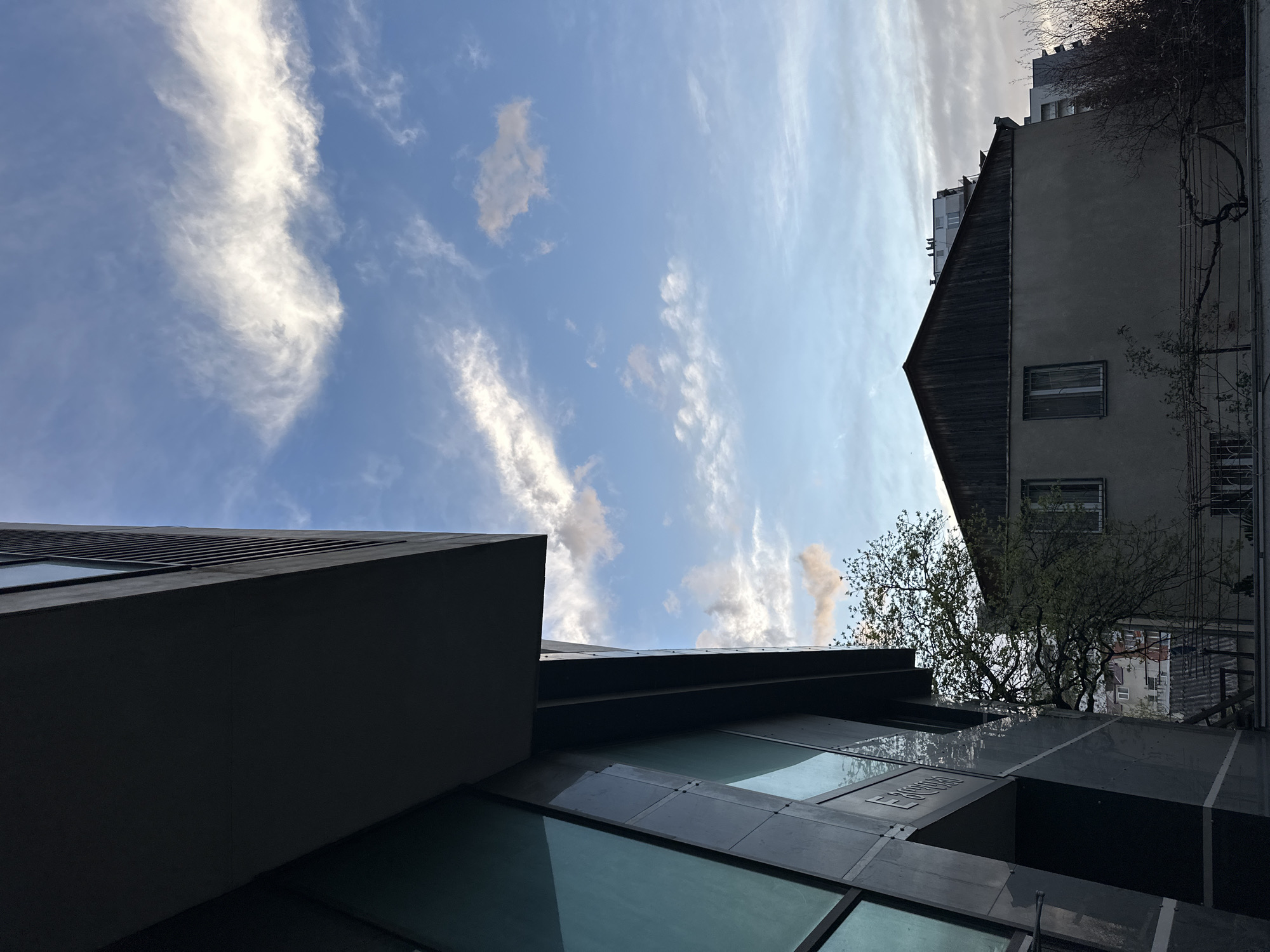 The image, captured from a low-angle perspective, showcases a vibrant blue sky with scattered white clouds, framed by two contrasting buildings and a tree, suggesting an upward gaze in an urban environment.
On the left, a modern building with dark, angular architecture dominates the foreground. Its facade is composed of dark, possibly metallic, panels and large glass sections that reflect the sky and other surroundings. Faint, partially legible text, appearing to spell "ERESKIE" or similar, is visible on one of the reflective glass surfaces.
To the right, an older building features a lighter-colored, possibly grey or beige, stucco facade and a distinctive dark, sloped roof. Several windows with grilles are visible on its side. In the background, behind this older structure, parts of a taller, lighter-colored apartment building are visible against the sky.
A tree with green foliage is situated between the two buildings, its branches reaching upwards towards the sky and partially obscuring the older building. The sky is bright blue, filled with various white clouds, ranging from thin, wispy streaks to more substantial, fluffy formations, indicating clear and fair weather. The bright illumination suggests it is daytime, likely morning or afternoon. The scene is static, with no discernible activity or people present. The architectural mix is characteristic of an urban setting like Tbilisi, Georgia.