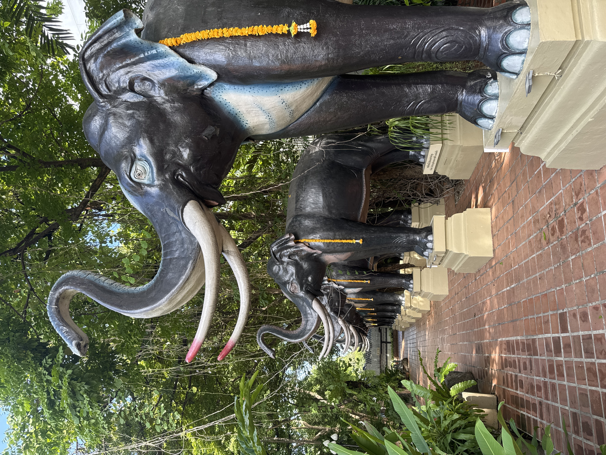 The image shows a row of black elephant statues with white tusks, each adorned with a yellow flower garland. The elephants stand on a low, light-colored wall or platform alongside a brick pathway. Lush green foliage surrounds the elephants, with trees visible in the background. The location appears to be outdoors. The presence of Thai script on a sign suggests the scene is in Thailand. Based on the query location, this is likely in Samrong Nuea Subdistrict Municipality.