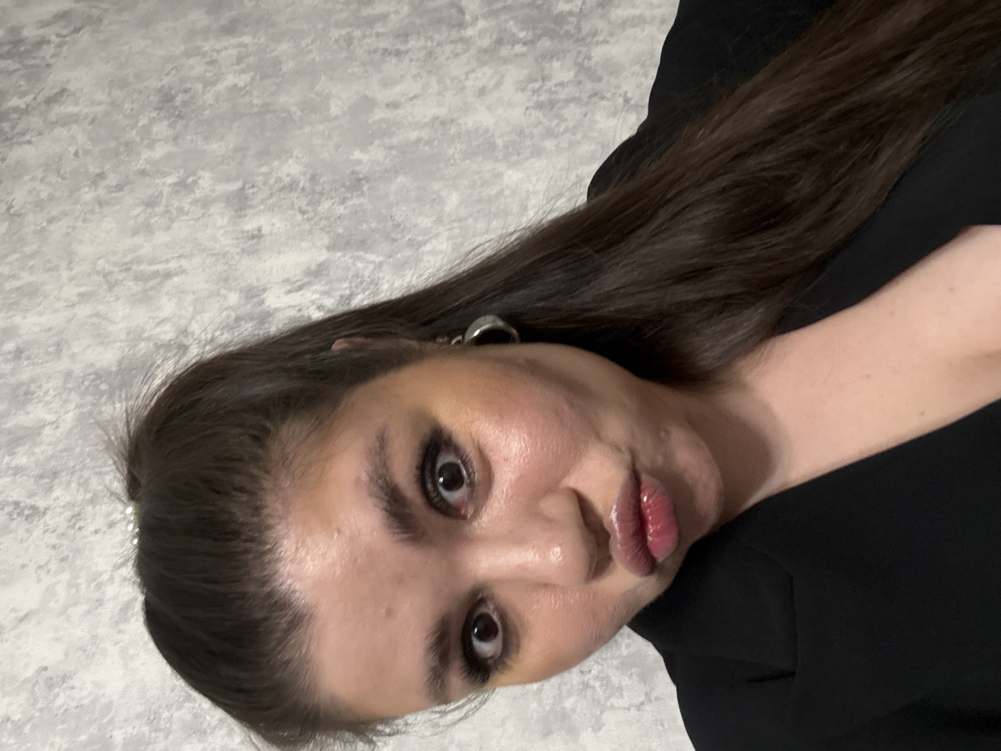 A woman is centered in the image, angled slightly. She has dark hair, fair skin, and is wearing dark clothing. The background appears to be a gray textured wall. The location is unknown.