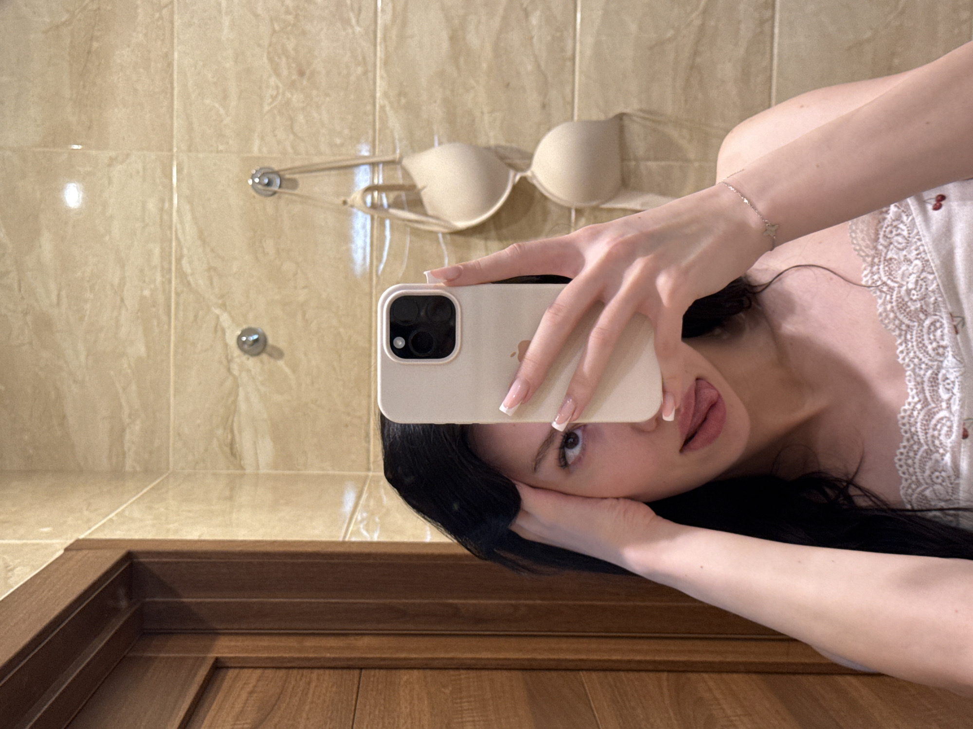 A young woman with dark hair is taking a mirror selfie in a bathroom. She is wearing a camisole with cherry details and a white lace trim. Her left hand is touching her head, and her right hand is holding a smartphone, obscuring part of her face. She is sticking her tongue out. In the background, there is a wall tiled with beige marble, two hooks, and a nude bra hanging on one of the hooks. A wooden door frame is visible on the left side of the image. The scene appears to be in a residential bathroom, possibly in the city of Khimki, Russia.