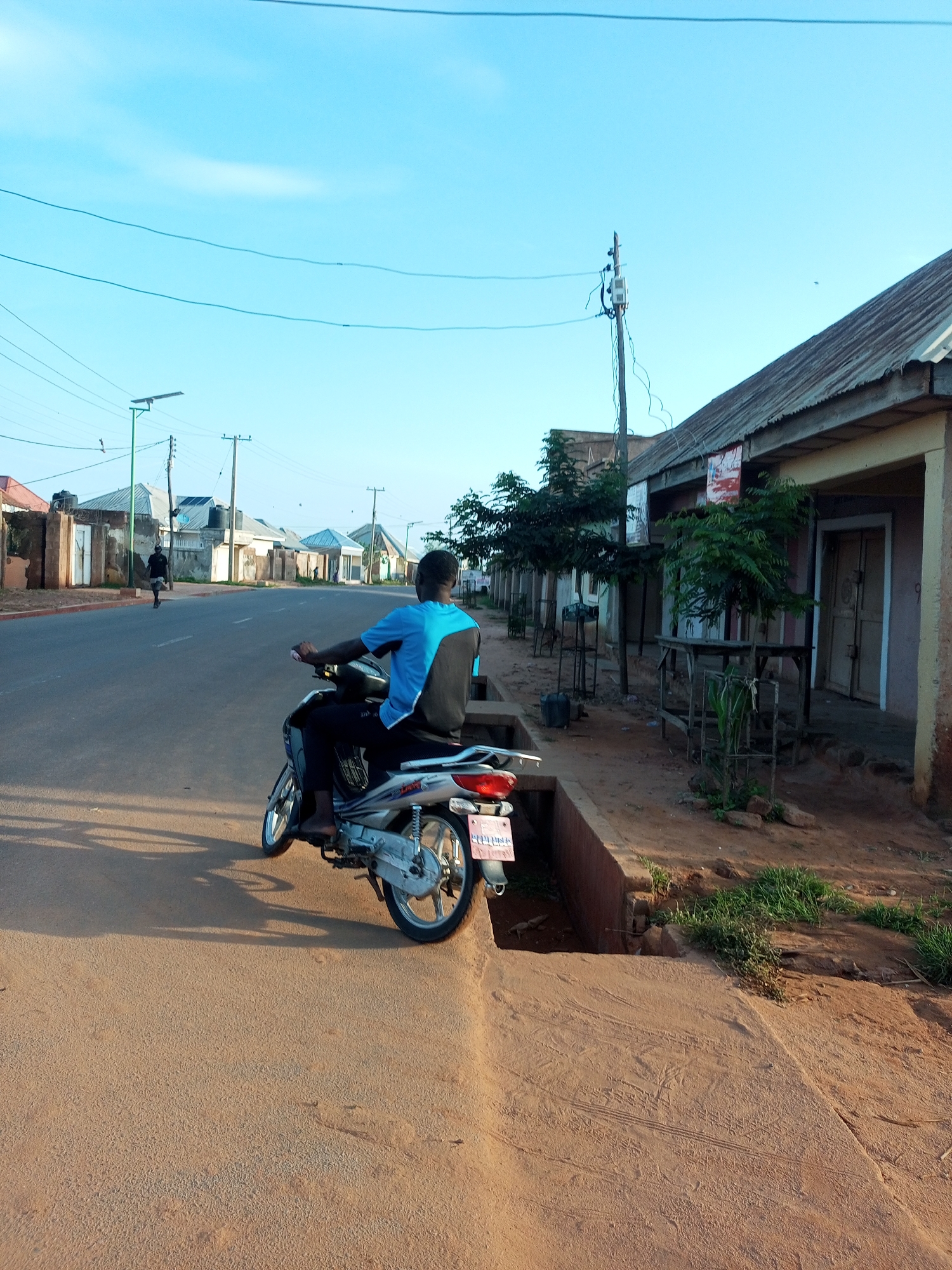 An outdoor, eye-level, medium shot captures a person riding a motorcycle on a paved road in Gombe, Nigeria, with buildings lining the right side of the street. The sky is a clear, light blue with faint wispy clouds. The road is mostly gray asphalt, with a sandy-colored shoulder on the right. On the left, another person is walking away from the camera.
The motorcycle rider is a young man wearing a blue and black t-shirt and dark pants. He is facing away from the camera, looking ahead down the road. His right hand is on the handlebar, and his left hand is resting on his lap. The motorcycle is silver and black, with a license plate clearly visible at the back.
The buildings on the right are single-story structures with corrugated metal roofs and entrances that appear to be doors. Some of the buildings have signs or posters on their facades. There are trees planted along the sidewalk in front of some of the buildings, and some of them are enclosed by metal cages. A drainage ditch runs along the edge of the sidewalk.
On the left side of the road, there are more buildings, some with what appear to be water tanks on their roofs. Telephone poles and streetlights are visible in the background, with power lines crisscrossing the sky. The overall impression is of a typical street scene in a populated area.