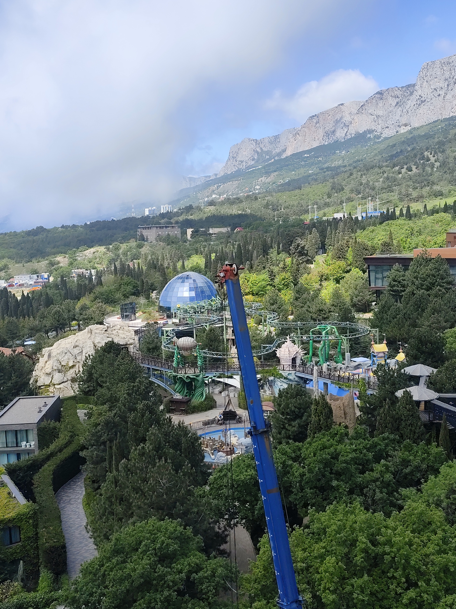 View from an elevated perspective in Симеизский поселковый совет, Ukraine, showing forested area, a mountain range in the distance, and a theme park. The park features a blue dome-shaped structure, and a green roller coaster. A blue crane is in the foreground obscuring some of the park. There are several other buildings and structures interspersed throughout the trees, and clouds cover parts of the sky and mountain range.