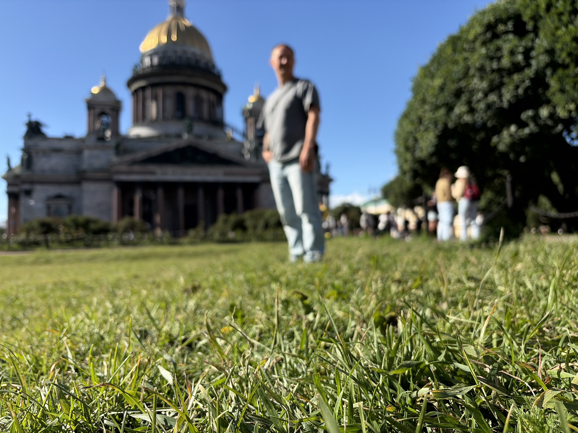 This media file depicts a sunny day in a park-like setting with a prominent domed building in the background. The focus of the image is the lush green grass in the foreground, shot at a low angle.
In the middle ground, a man is standing, facing the camera. He is wearing a grey t-shirt and light-colored jeans, with his hands in his pockets. He appears relaxed. Behind him, other people are visible, some walking and some standing, suggesting a public space with visitors. The domed building, likely a cathedral, is grand and features classical architecture with columns and ornate details. Its golden dome gleams in the sunlight. To the right of the building, a large, dense green tree provides shade.
The clear blue sky indicates fair weather. The bright sunlight suggests it is likely daytime, possibly late morning or afternoon. The presence of tourists and the architectural style of the building strongly suggest the location is Saint Petersburg, Russia, specifically near Saint Isaac's Cathedral. No text is visible in the image.