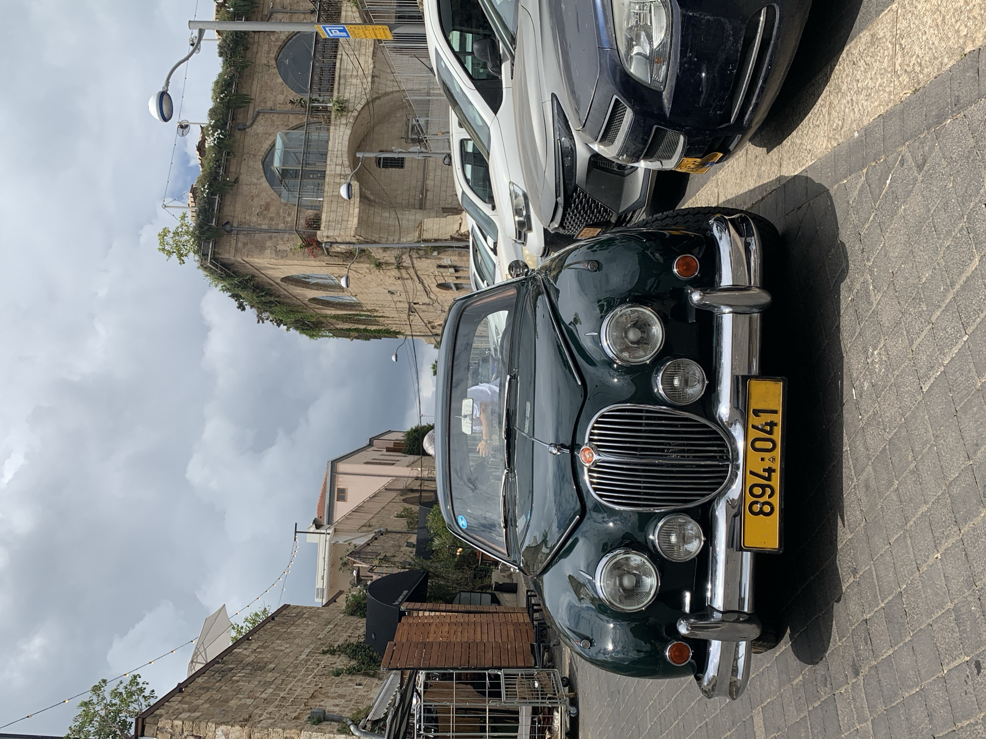 The image shows the front of a dark green vintage Jaguar car parked on a cobblestone street in what appears to be an older neighborhood. The car has a yellow Israeli license plate with the number "894-041". Behind the Jaguar, parts of two modern vehicles (one white and one dark) are visible.
The background includes buildings of varying architectural styles, some appearing to be older structures with arched windows and stone facades. Vegetation, including plants and trees, is visible on some of the buildings. A street lamp and overhead string lights are also present. The sky is overcast.
The location is likely in the city of Tel Aviv, Israel, given the license plate on the vehicle.