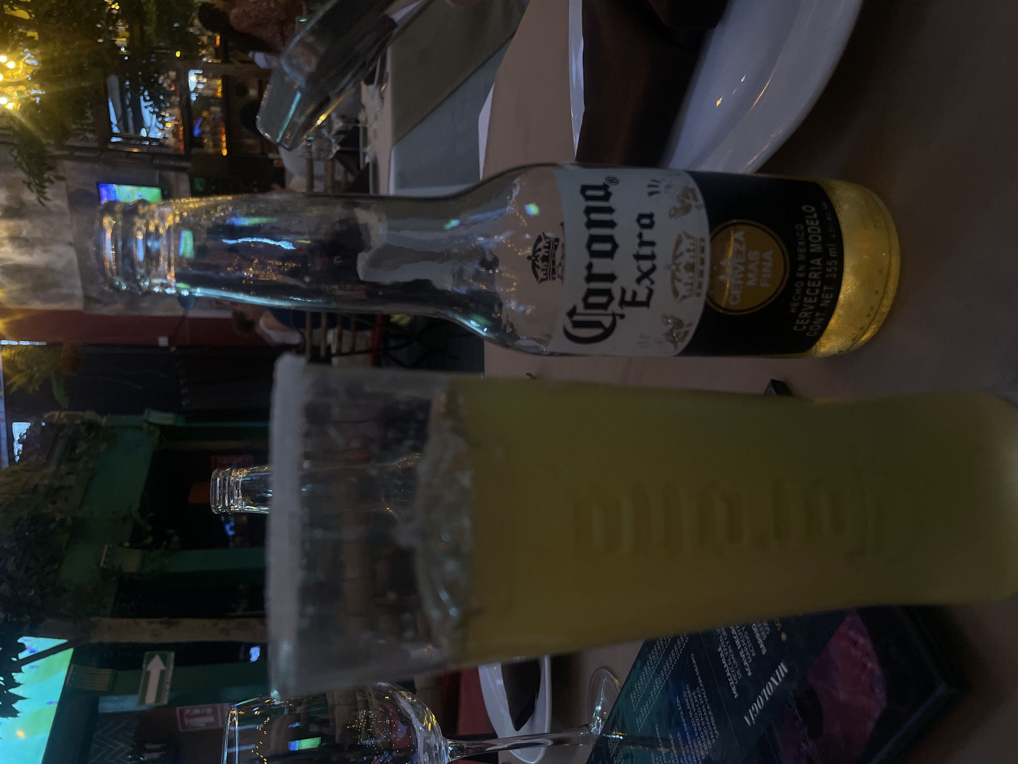 The image shows a partially consumed bottle of Corona Extra beer and a margarita with a salted rim, likely on a table at an outdoor dining area in Puebla, Mexico. The bottle label is visible, indicating the brand and origin of the beer. The menu for "Mixologia" and other drinks are also visible on the table. The background reveals a restaurant or bar setting with people and ambient lighting.