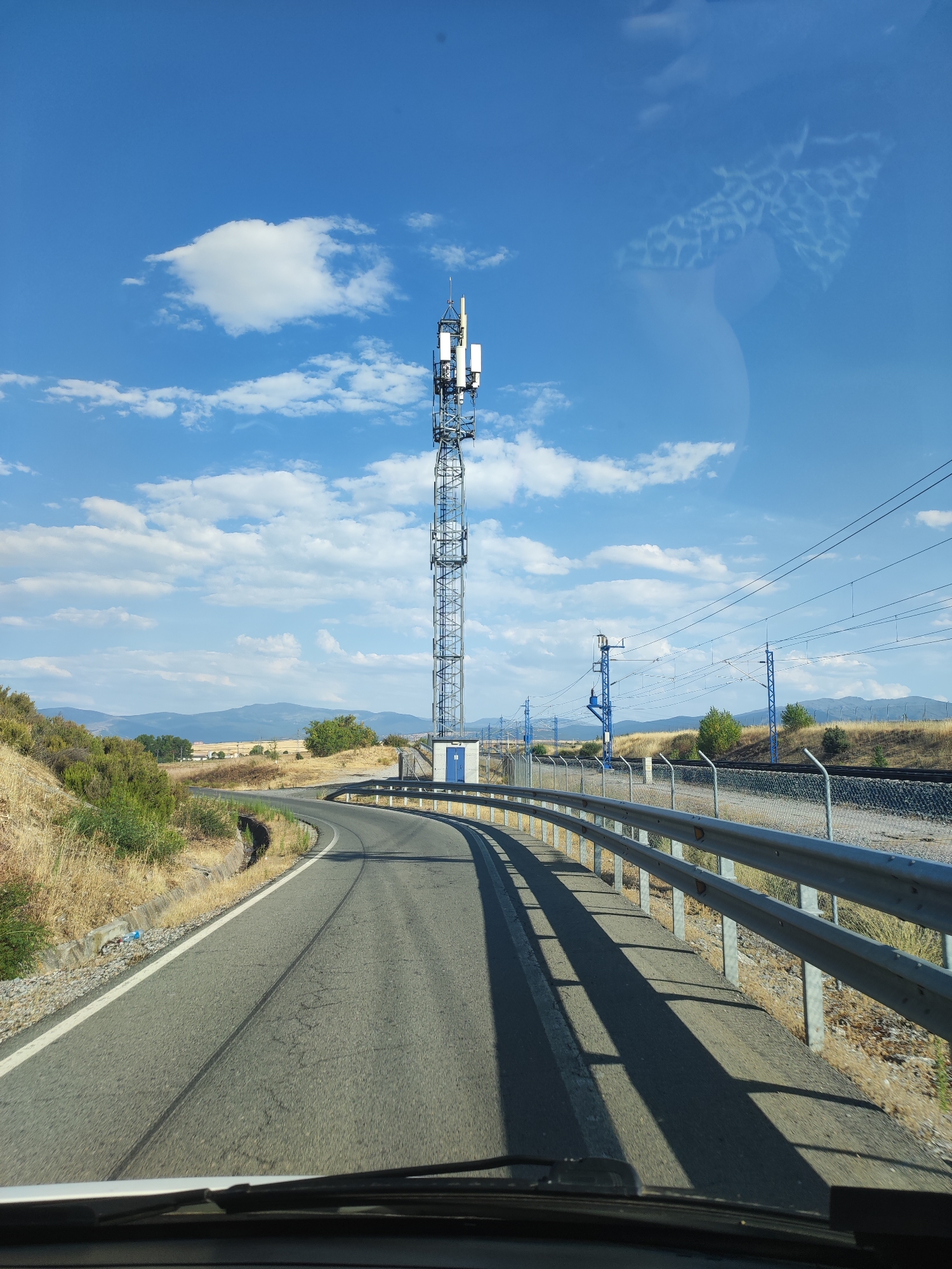 The image shows a view from a vehicle driving on a road. The road curves to the left and is bordered by a metal guardrail on the right. A cell tower with multiple antennas is visible to the left of the road. Adjacent to the cell tower is a small blue building. Beyond the guardrail, train tracks run parallel to the road, with overhead power lines supported by blue poles. The landscape is dry and rural, with sparse vegetation, hills in the distance, and a bright blue sky with scattered white clouds. There is a reflection on the windshield, possibly of a patterned object. The scene appears to be located in Valverde del Majano, Spain, near the train tracks.