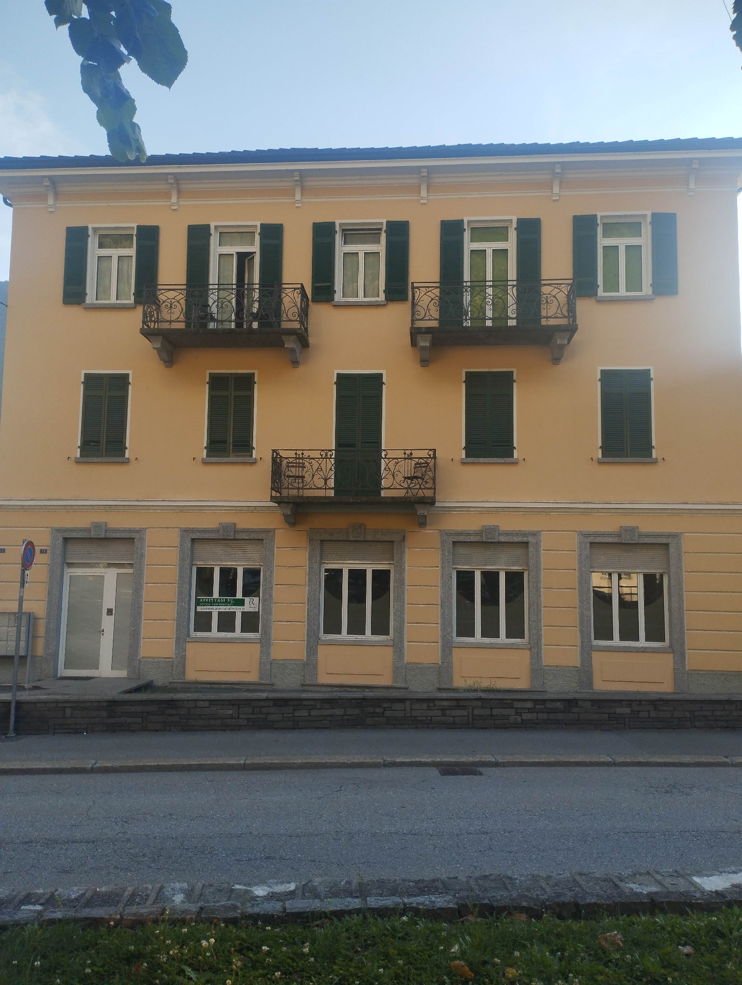 A multi-story building with a light yellow-orange facade stands front and center on a paved street in Biasca, Switzerland. The building features a dark blue tiled roof with a white cornice detailed with corbels.
The upper two floors display a consistent pattern of windows with dark green louvered shutters. On the uppermost floor, there are three sets of double windows. The leftmost window has closed shutters. The central window is open inwards, its green shutters swung wide, and it is fronted by an ornate wrought-iron balcony with scrollwork designs, supported by two grey stone corbels. The rightmost window also has its green shutters open.
Below this, on the intermediate floor, there are three single windows, each with closed dark green shutters. Centered below the main balcony and between the windows of this intermediate floor, a smaller, narrower wrought-iron balcony with similar decorative elements is affixed to the facade.
The ground floor of the building is distinguished by a grey stone base and grey stone framing around its openings. From left to right, this floor includes: a white door with a small frosted glass panel, positioned next to a closed grey roller shutter; a window with white frames, covered by a closed grey roller shutter above, displaying a green and white "AFFITTASI" (For Rent) sign with contact information and an "R" logo; and another window with white frames, also covered by a closed grey roller shutter above it. The sections of the facade between the grey stone frames on the ground floor match the yellow-orange color of the upper stories.
In the foreground, a grey asphalt road with minor cracks stretches across the width of the image, separated from the building by a grey concrete curb and a narrow strip of pavement. Immediately in front of the road is a strip of green grass dotted with small white clover flowers and a few scattered brown leaves. To the far left, a red and blue circular road sign, likely indicating "no parking" or "no stopping," is visible on a grey pole. The sky above is clear and light blue, with dark green leaves from an unseen tree partially entering the frame from the top left. No people or animals are present in the scene.