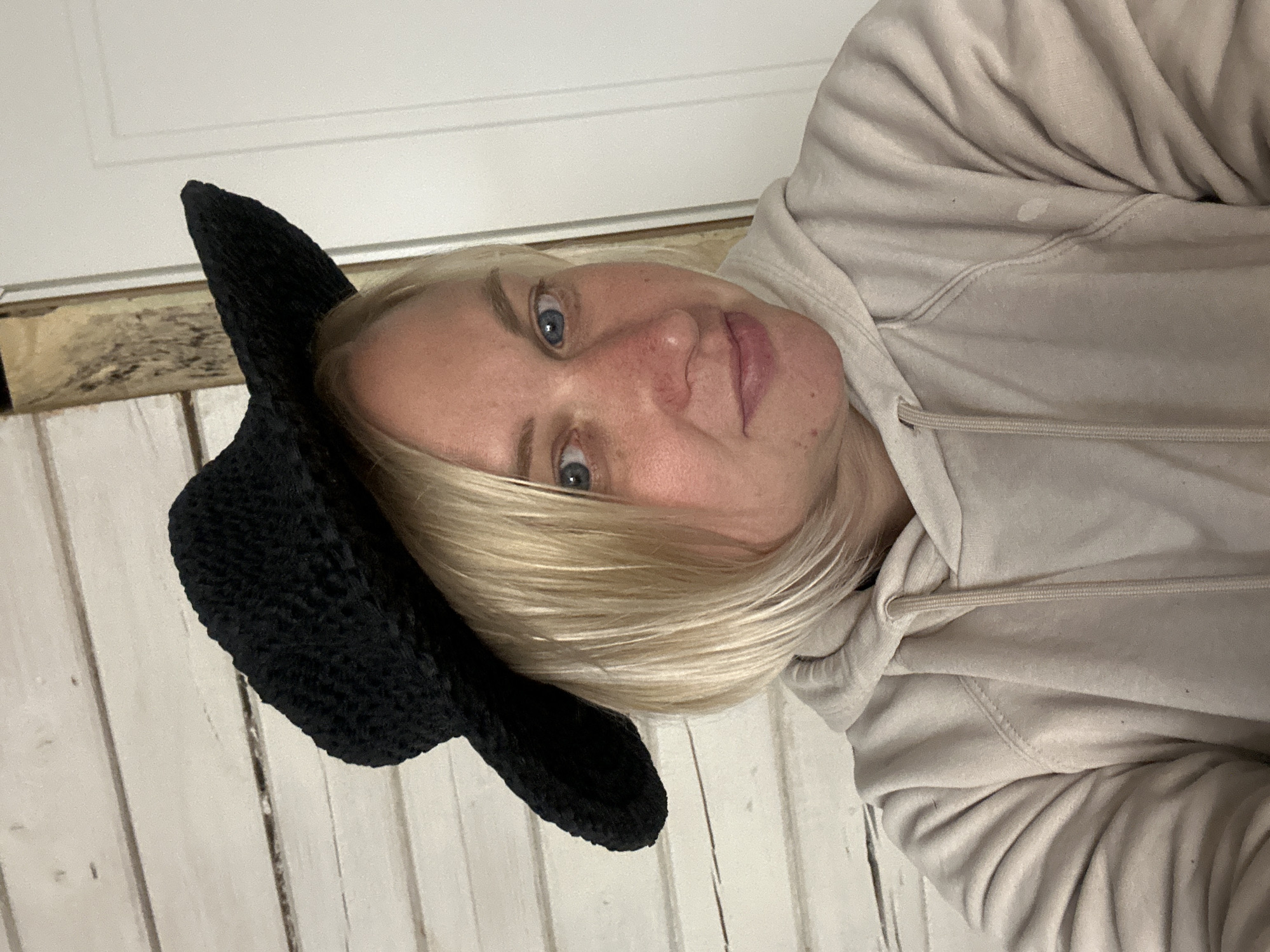 A blonde woman with blue eyes wears a black crochet cowboy hat and a beige hooded sweatshirt. She is indoors, with a white wooden plank wall behind her. A vertical wooden beam is visible to her right. The lighting is diffused, suggesting an indoor setting. The scene does not contain any contextual information indicating a specific location within the city of Chaikovsky, Russia.