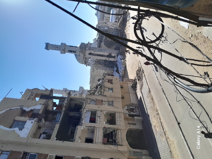 The image captures a scene of extensive destruction in an urban environment, likely in Khan Yunis, Palestinian Territory. In the foreground, several multi-story buildings are severely damaged, with large sections of their external walls and interiors blown out, revealing mangled rooms, debris, and structural elements. The extent of the damage suggests the aftermath of bombings or heavy shelling.
In the mid-ground, a mosque with a distinctive minaret and a dome is visible under a clear blue sky. While appearing somewhat less damaged than the immediate foreground structures, it also shows signs of impact or wear. A paved road, flanked by dusty, rubble-strewn ground, runs through the scene. Overhead, numerous tangled power lines and cables crisscross the frame, some appearing thick and coiled, further emphasizing the disarray.
The scene is illuminated by bright daylight, with clear skies and shadows indicating either morning or afternoon. There are no people visible in the frame. The overall impression conveyed is one of profound devastation and the grim reality of conflict. In the bottom right corner of the image, the watermark "Galaxy A14" is visible, indicating the device used to capture the photograph.