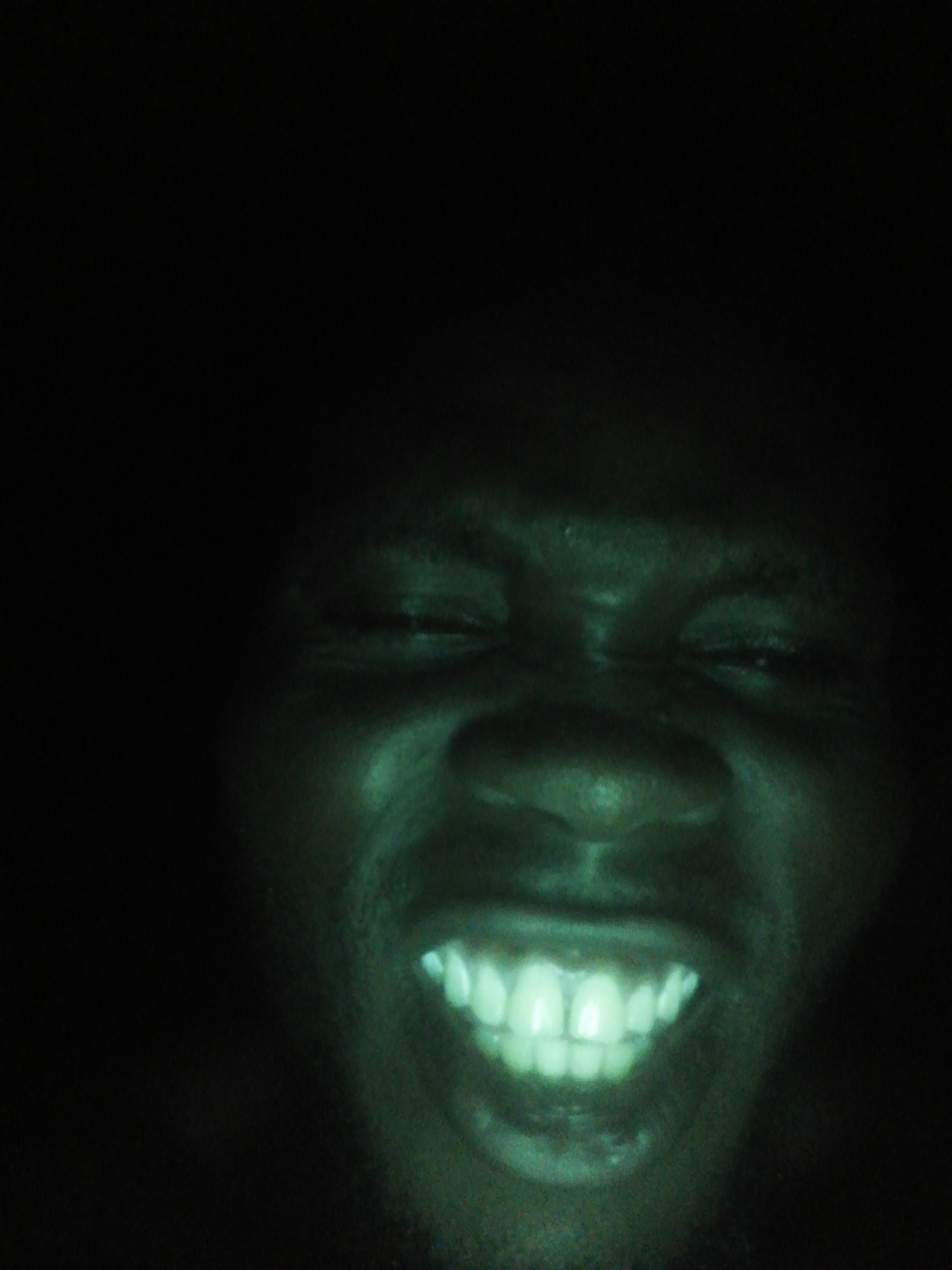 A dark, close-up image shows a person's face, illuminated with a green tint. The person is smiling, revealing their teeth. The background is entirely black. The location is unknown.
