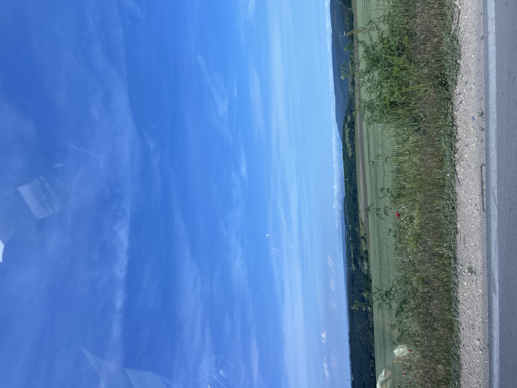 The image presents a wide-angle landscape captured on a bright, clear day, likely from a moving vehicle given the reflections visible on the left side of the frame (which would be the top when oriented correctly).
When viewed horizontally, the scene is dominated by a vast, vibrant blue sky filling the upper two-thirds of the image, featuring only a few faint, wispy white clouds. Below the sky, a distant horizon line reveals a range of undulating hills or mountains, possibly covered in trees or distant settlements, stretching across the width of the photograph.
The lower third of the image is occupied by expansive green fields, suggesting agricultural land, extending from the midground towards the distant hills. In the foreground, a grassy verge with various plants and weeds runs alongside a paved road, which occupies the far right edge of the frame. Several vibrant red poppies are scattered within this green verge and the adjacent field. The road is light gray with a clear white line marking its edge.
No specific human activity or people are visible. The time of day appears to be midday due to the bright, even lighting and lack of prominent shadows. The weather is sunny and clear. No text is visible in the image. This serene, open rural landscape is situated near Kichevo, Bulgaria.