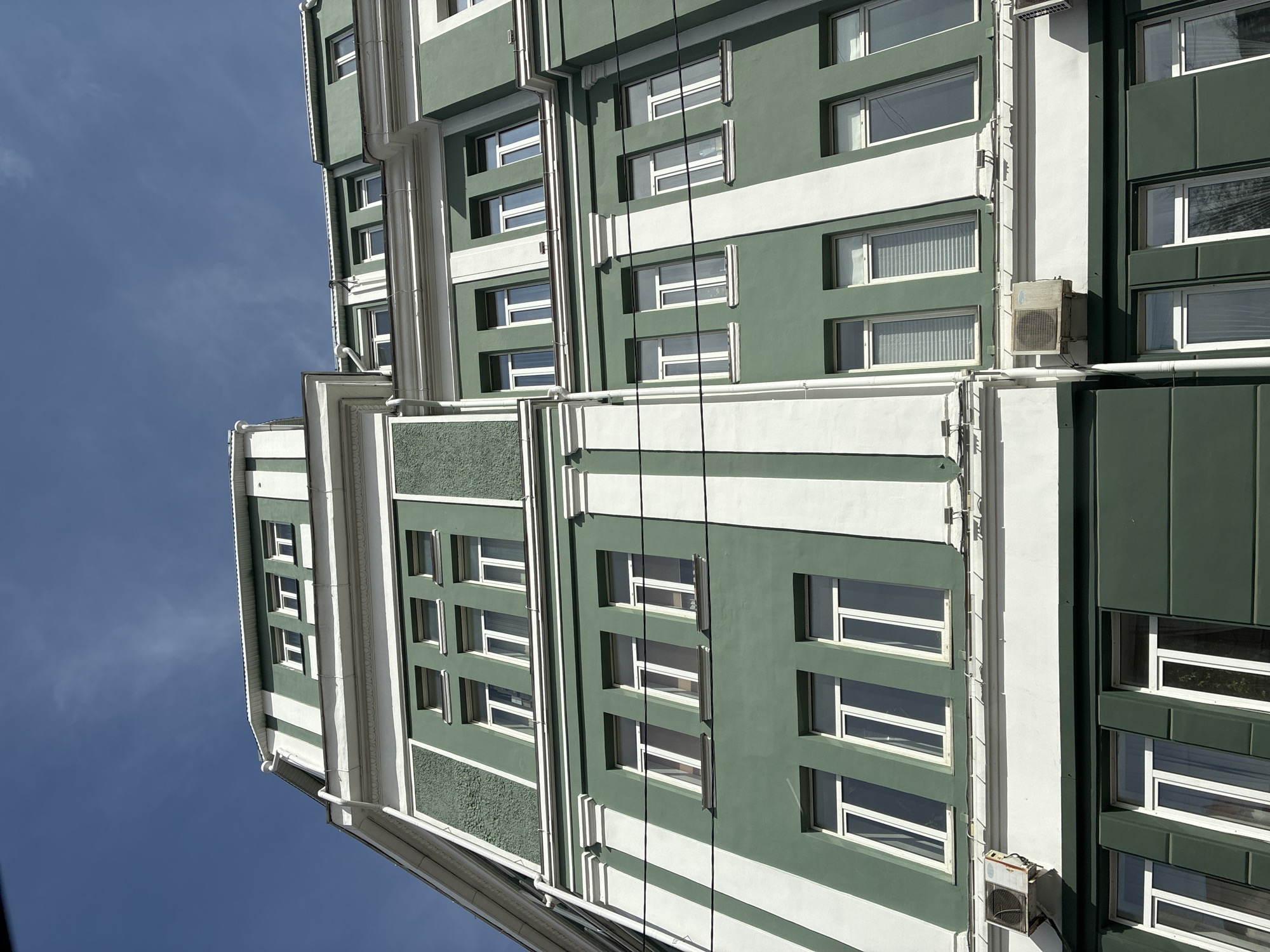 The image shows the exterior of a building in an unknown city. The building has a green and white color scheme with multiple windows. There are wires running vertically along the facade of the building. The sky is blue and mostly clear. The location and other details are unknown.
