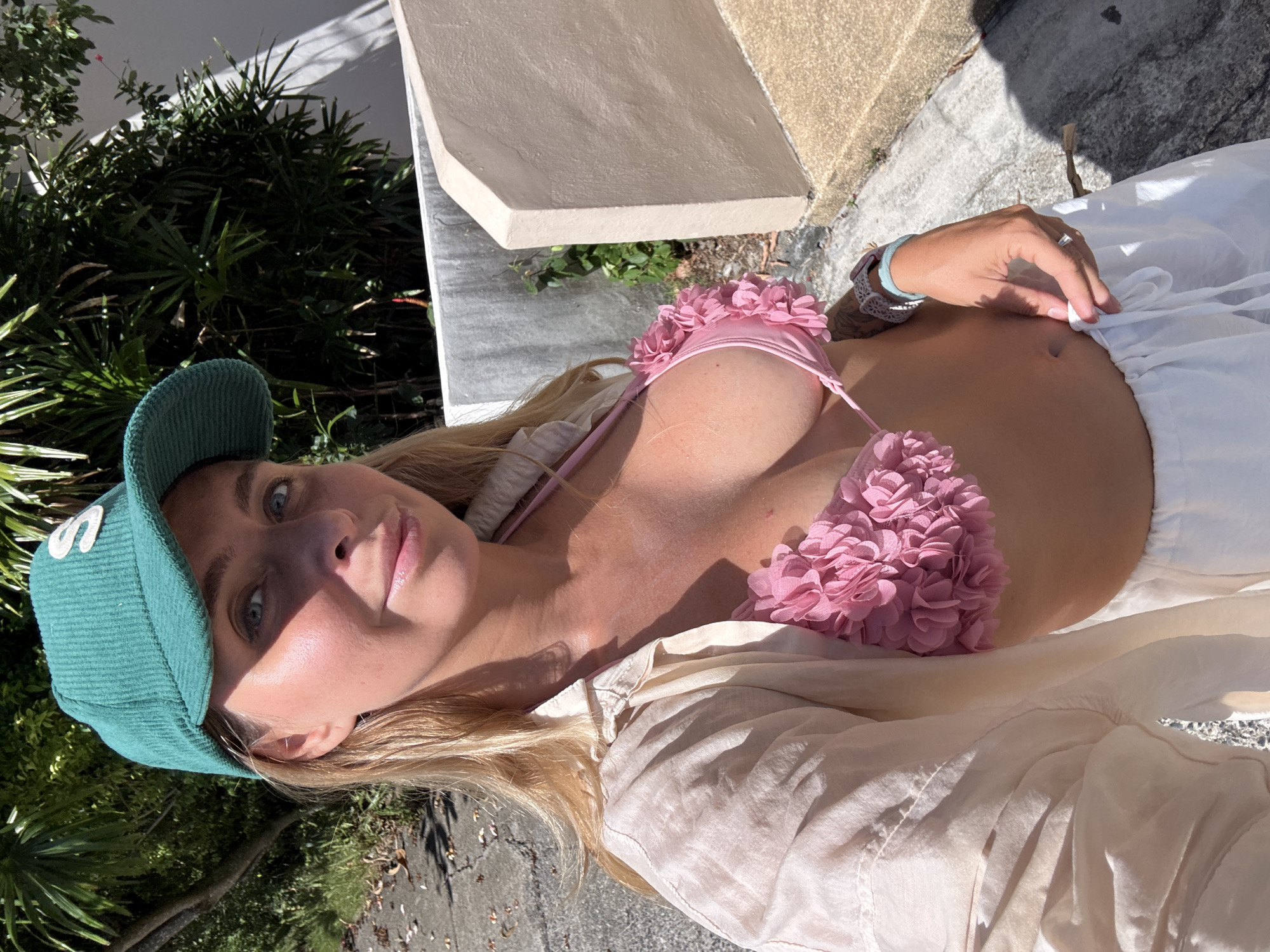 A woman wearing a green cap and a pink floral bikini top is pictured outdoors in Ko Samui District, Thailand. She is also wearing a light-colored button-down shirt and white pants. The background consists of lush green foliage and a concrete structure. The woman is taking a selfie, with the sun casting shadows on her face and body.