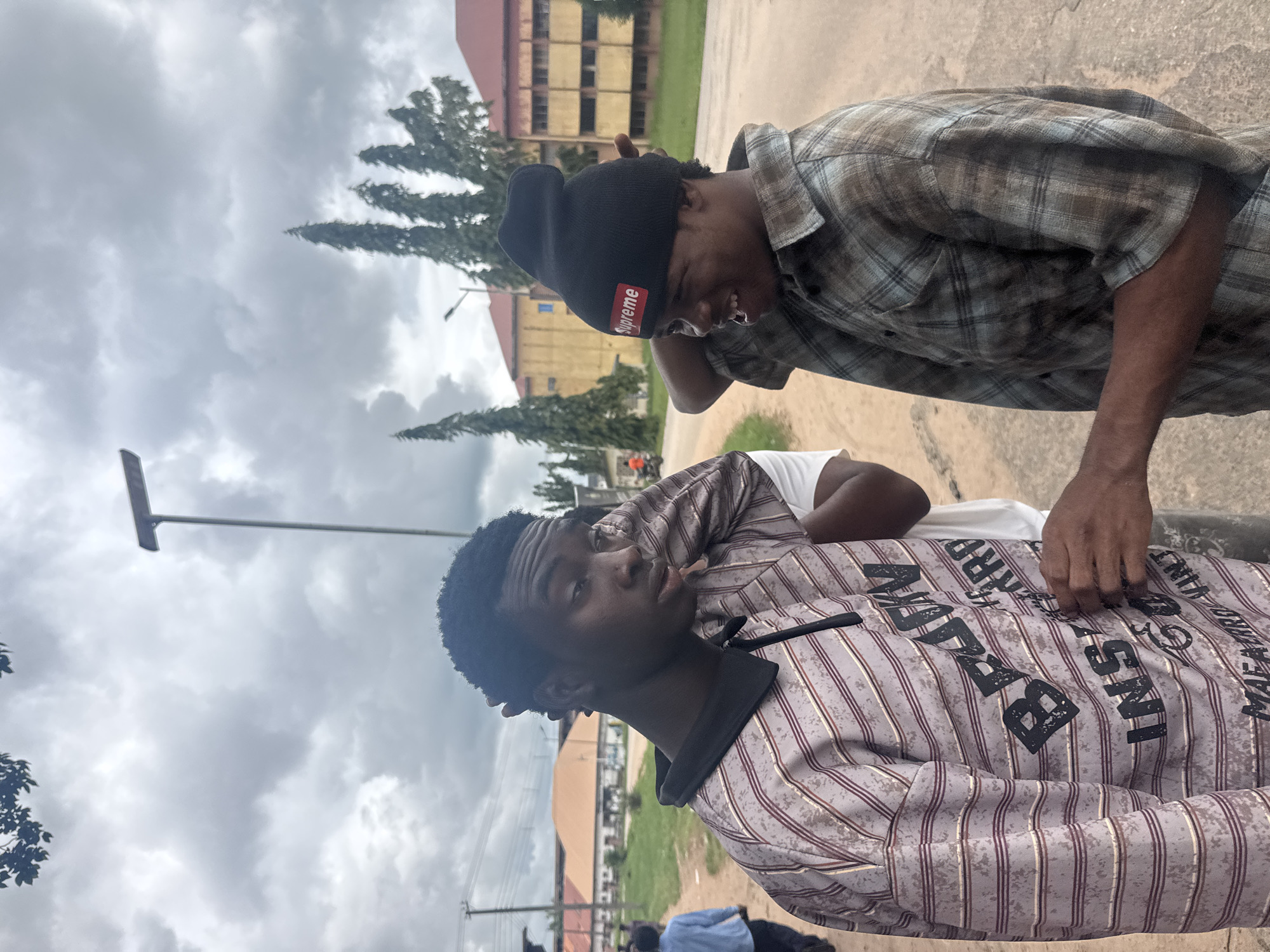 Two young Black males are standing on a paved surface in what appears to be a city environment. The male on the left is wearing a striped long-sleeved collared shirt with text printed on it. He is looking to his right, touching his temple with his right hand. The male on the right is wearing a plaid long-sleeved shirt and a black beanie with a red "Supreme" logo. He is looking to his left and speaking, with his mouth open. His left hand is raised and extended towards the other male. In the background, there are buildings, trees, a street lamp, and other indistinct figures. The sky is overcast with clouds. The location is identified as Ihiagwa, Nigeria.