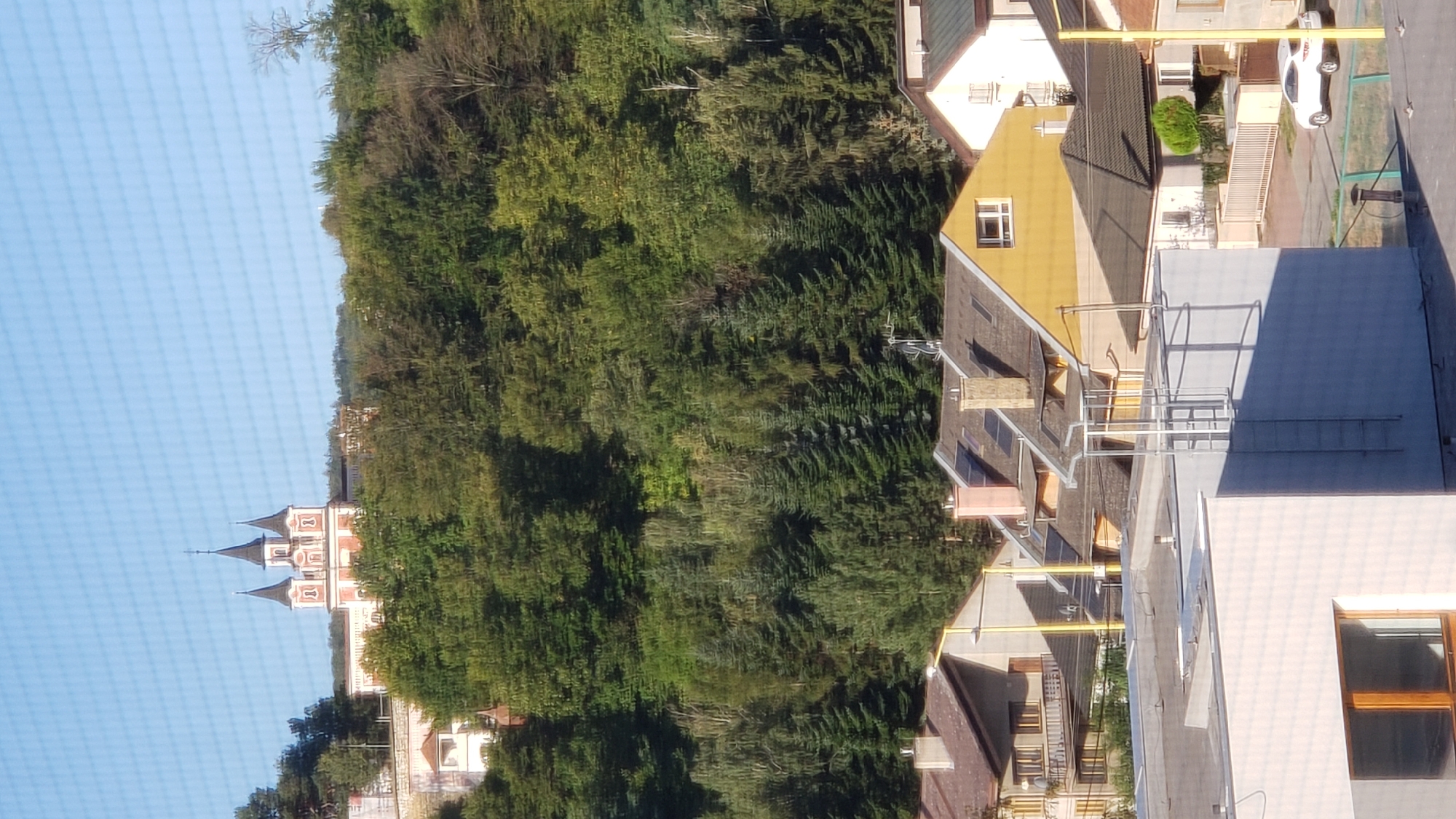 This is an eye-level, medium shot of a residential area in Prešov, Slovakia. The foreground features a modern, light-colored building with a large window. Behind this building, several residential houses with pitched roofs are visible. The roofs are made of various materials, including shingles and tiles, in colors such as brown, yellow, and gray. Some houses have chimneys and dormer windows. A white car is parked on the street in front of one of the houses.
In the middle ground, a dense forest of deciduous and coniferous trees covers a hillside. The trees vary in color from deep green to lighter shades, suggesting a mix of species and possibly the season.
In the background, on top of the hill, a distinctive pink and white building with three spires stands out against the clear blue sky. This appears to be a church or a similar religious institution. The sky is bright and cloudless, indicating fair weather. The overall scene depicts a suburban or semi-rural setting with a mix of residential architecture and natural landscape, with a prominent landmark on the hill.