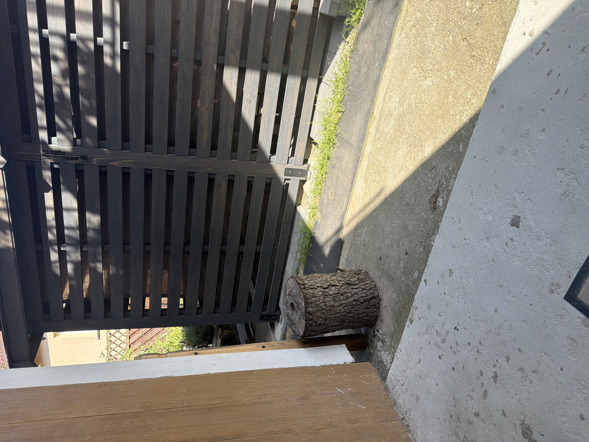 The image depicts an outdoor scene featuring a rustic wooden stump positioned on a concrete surface next to a wooden plank wall. Behind the stump, a dark wooden slatted fence or gate is visible, with sunlight casting shadows across its surface. The environment appears to be an exterior space, possibly a yard or a pathway, with some grass growing along the edge of the fence. The overall impression is one of a somewhat unkempt but functional outdoor area.
