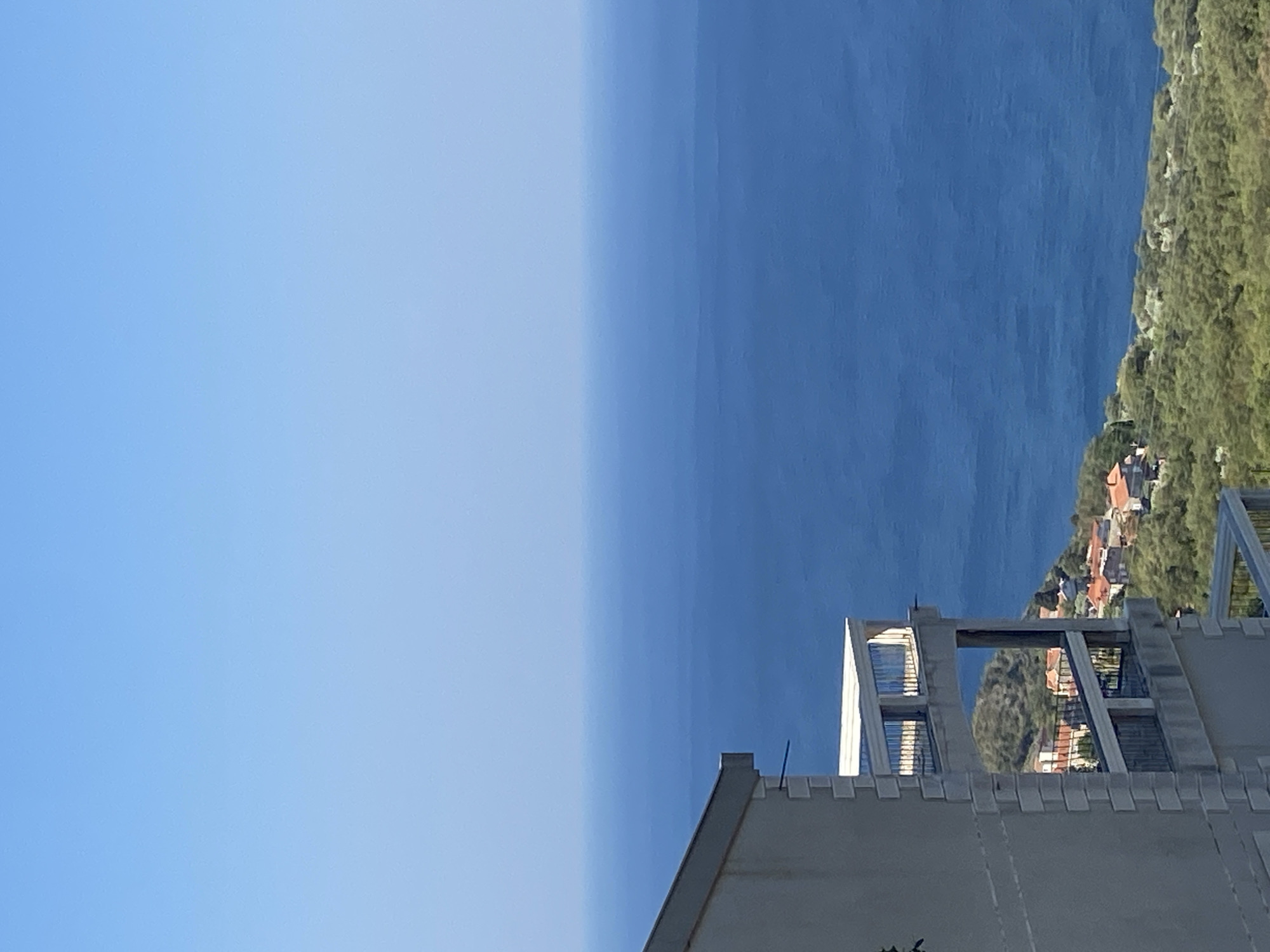 The image shows a view from a building in Rađenovići, Montenegro, overlooking the Adriatic Sea. The sky is a clear, pale blue, transitioning to a deeper blue at the horizon where it meets the sea. The sea itself is a vast expanse of deep blue water, with some subtle ripples visible on its surface. In the foreground, the side of a building is visible, constructed of light-colored stone with distinct horizontal lines of darker stone. This building has at least two levels of balconies, each with metal railings. One balcony, on the upper level, is partially visible and extends outwards. Below the building, a hillside is covered in dense green foliage and scattered with red-tiled roofs of houses, indicating a small village or settlement nestled into the landscape. The overall scene depicts a coastal, Mediterranean environment on a clear day.