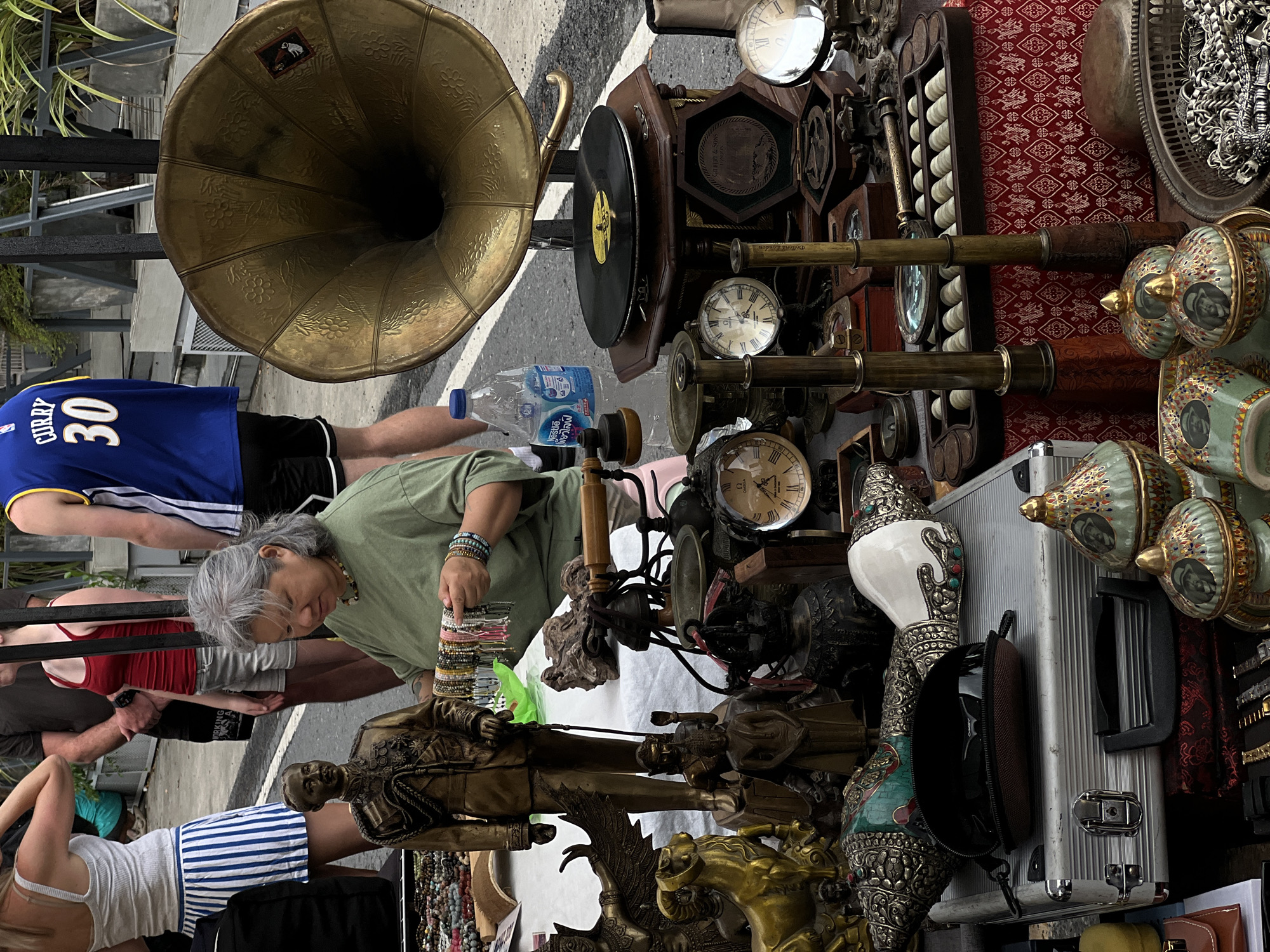 A woman with gray hair wearing a green shirt is examining bracelets at an outdoor market stall. She is standing behind a table displaying various items, including statues, clocks, a gramophone, and other decorative objects. In the background, several people are visible, including a person wearing a blue basketball jersey with "CURRY 30" on the back. The scene appears to be a street market.