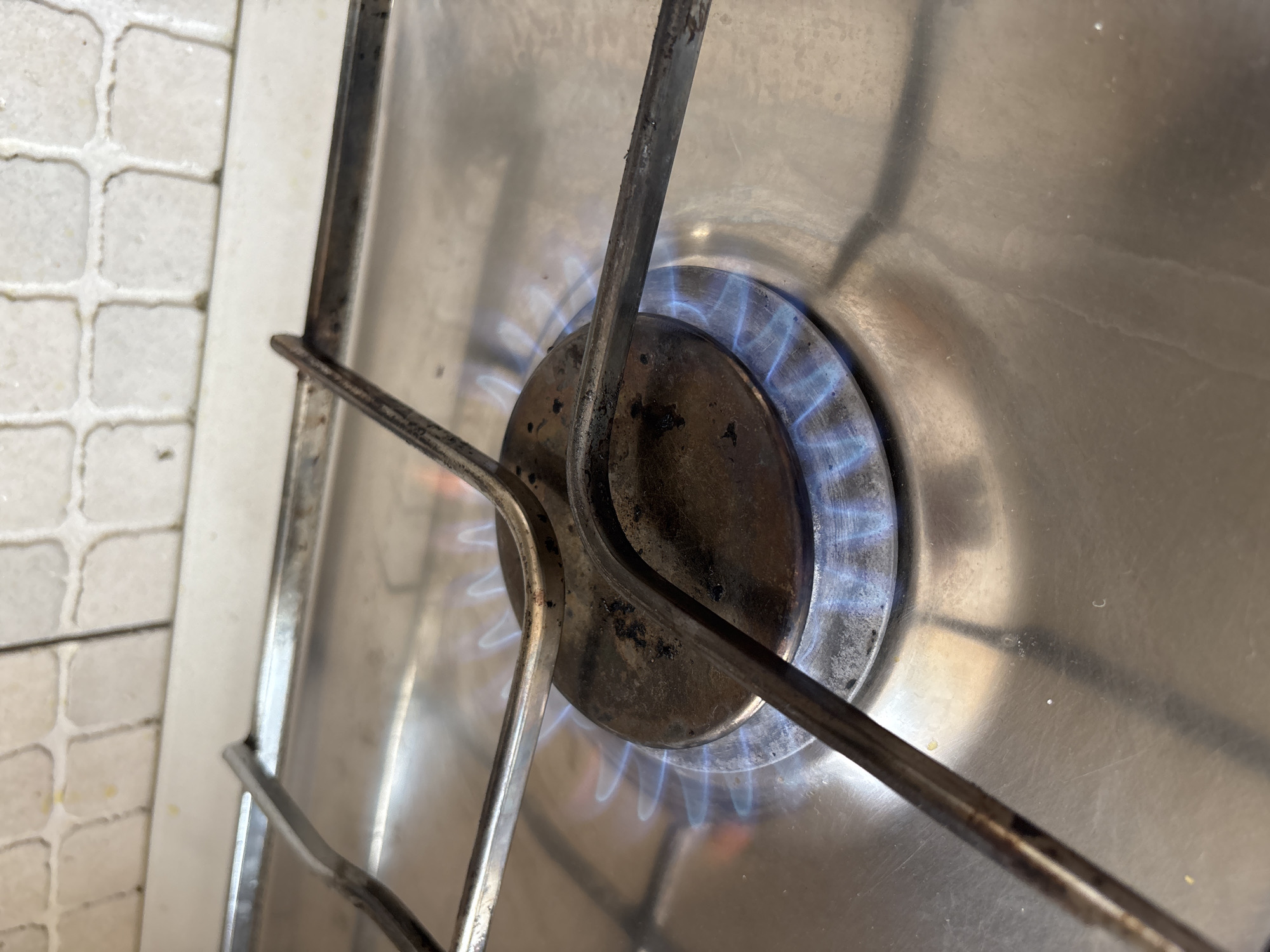 A close-up shot shows a gas stove burner with a blue flame. The burner is made of dark metal and appears slightly worn. Two metal grates are positioned over the burner, one in the foreground and one angled away. The stove surface is made of stainless steel and reflects light. To the left of the stove, a tiled backsplash with square, off-white tiles is visible. The tiles are separated by grout, creating a grid pattern. The scene suggests a kitchen environment, likely in a home. The lighting is bright, indicating daylight. The image is static, so no specific event is captured beyond the burner being on. No people, text, or weather conditions are discernible. The location context provided is Krasnodar, Russia.