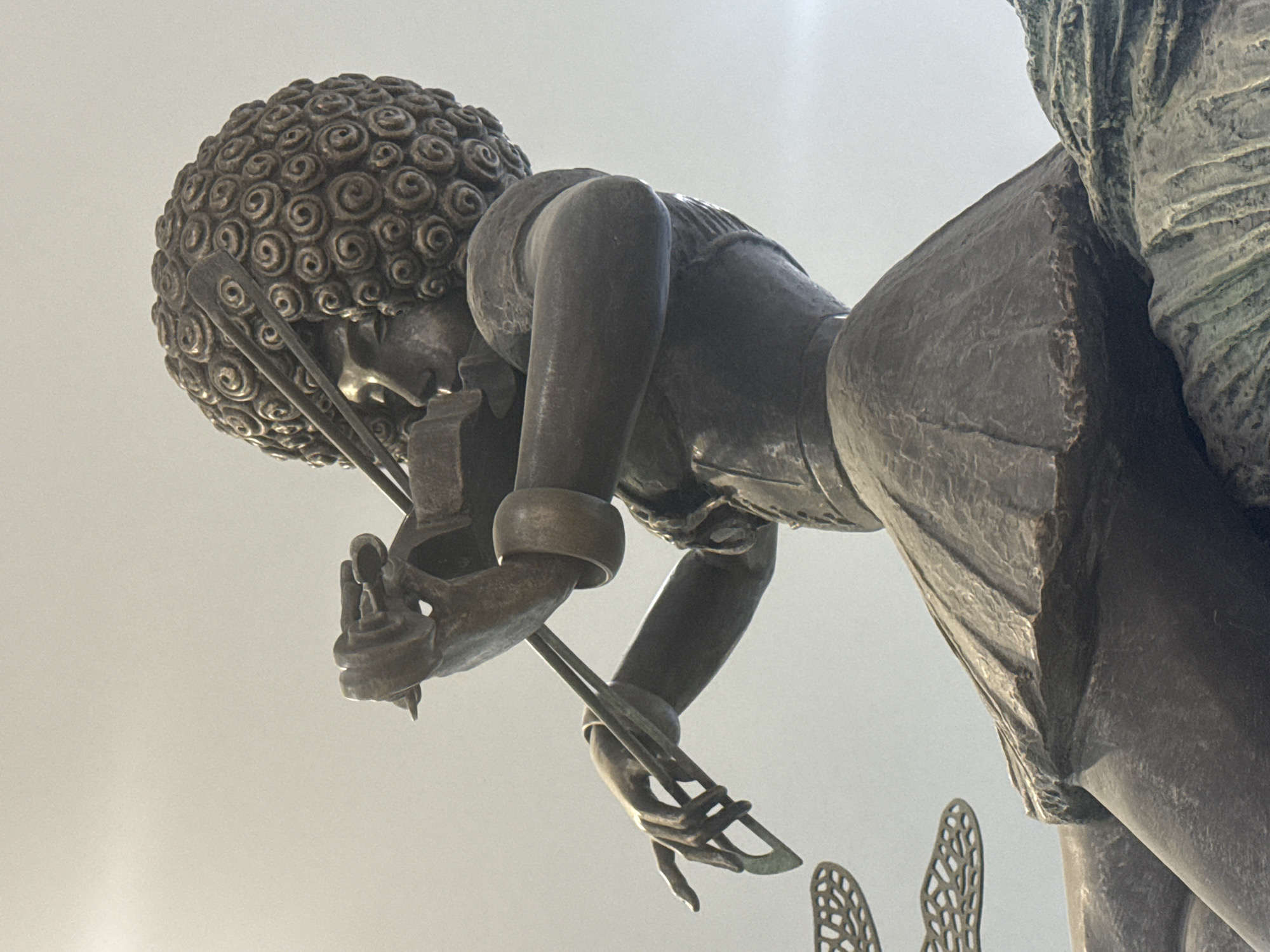 A low-angle shot captures a bronze statue of a woman, identifiable as one of the figures from the sculpture "Gold Mining Women" in Yekaterinburg, Russia. The statue is depicted with a curly afro hairstyle, wearing a garment, and is adorned with arm bracelets. The woman is bent over, and her hands are seen holding tools. In the background, a blurry gray sky is visible. The statue is viewed from below, emphasizing its scale and intricate details.