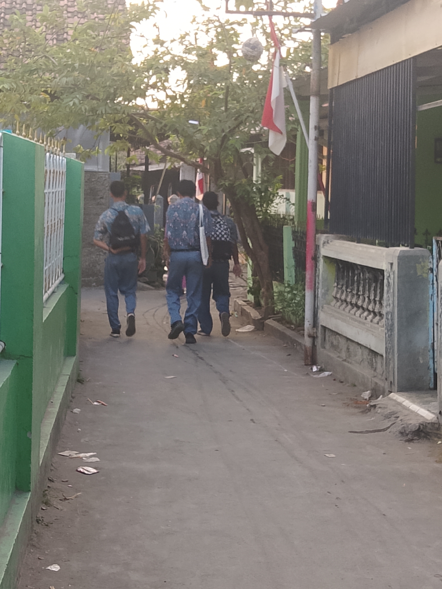 Three young males, appearing to be students wearing matching uniforms consisting of blue patterned shirts and blue pants, walk away from the camera down a narrow paved street. The two individuals in front are walking closely together, while the person on the left is slightly ahead. The male in the middle carries a black backpack. The male on the right carries a white tote bag. The street is lined with buildings on the right and a green fence on the left. A tree with green foliage is present on the left side of the street, and a tall pole with a red and white Indonesian flag is visible on the right. Debris, including paper scraps, is scattered on the street and along the sides. The lighting suggests it is either early morning or late afternoon. The architectural style of the buildings and the presence of the Indonesian flag suggest the location is Cirebon, Indonesia, likely in a residential area.