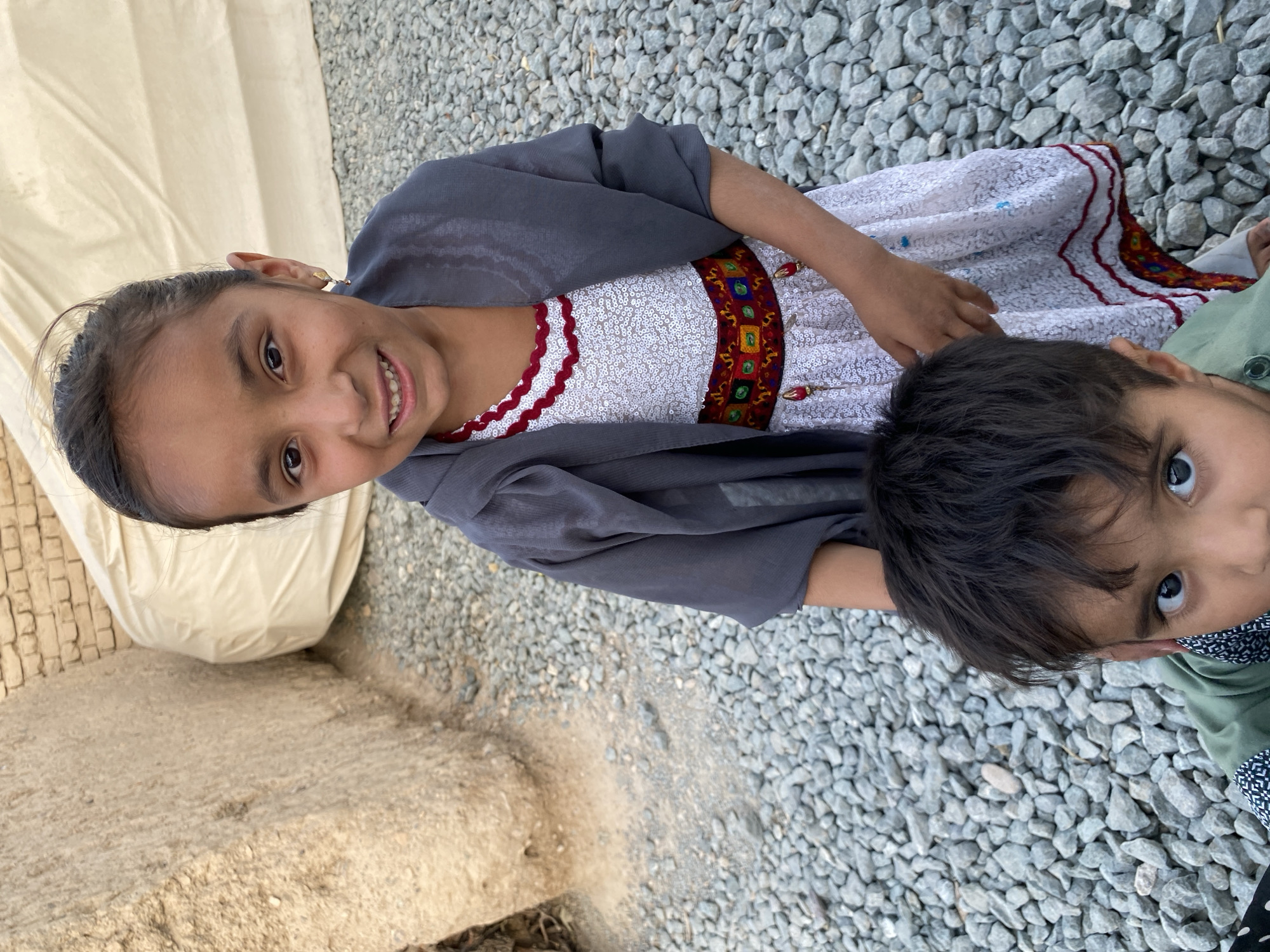 In the city of Kandahar, Afghanistan, two young children are pictured standing outdoors on a ground covered with grey gravel. The older child, a girl, stands in the foreground, facing the camera with a slight smile. She has dark hair pulled back and is wearing a white dress adorned with intricate red and gold embroidery, a red and white patterned trim around the neckline, and a sequined overlay. She has a grey shawl draped over her shoulders. To her left and slightly in front, a younger child, a boy, is partially visible, looking directly at the camera with wide, dark eyes. He has dark, tousled hair and is wearing a green shirt. In the background, a large, rough-textured, light brown structure is visible on the left, and a cream-colored, tent-like structure stretches across the upper background. The overall environment appears to be an outdoor, possibly rural or semi-constructed area.
