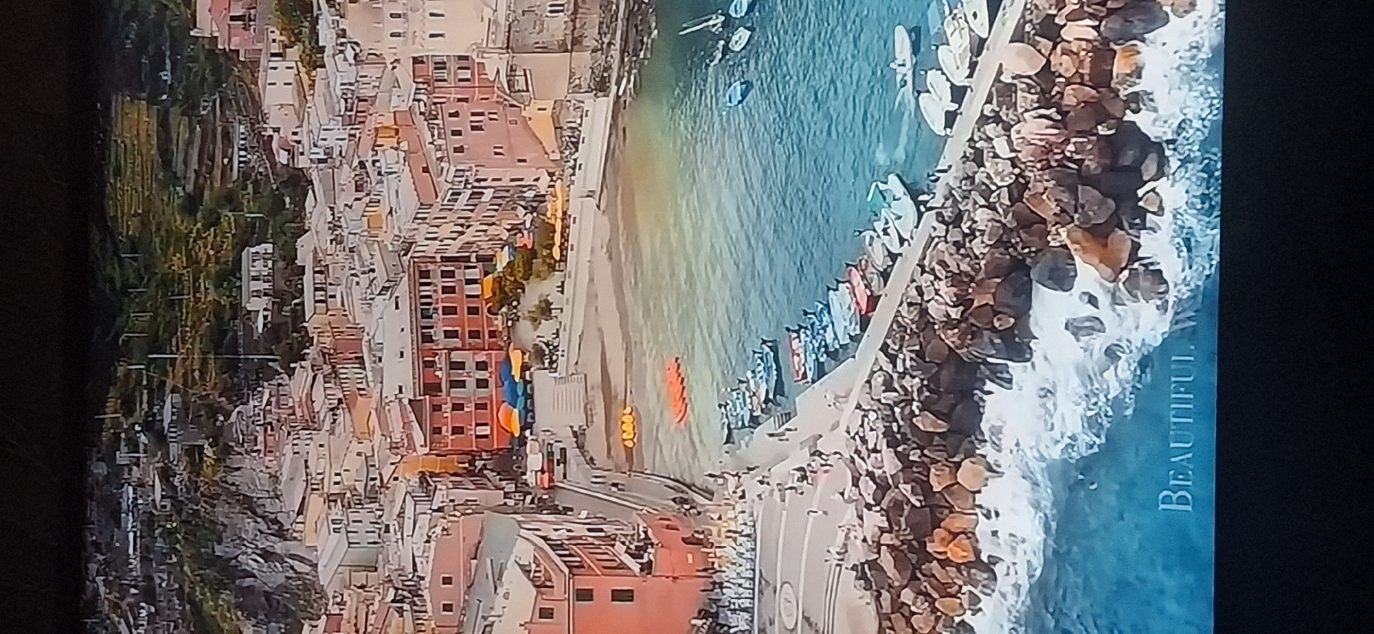 An aerial view of a coastal town shows colorful buildings cascading down a hillside towards a harbor filled with boats. A stone breakwater juts out into the turquoise water, where waves crash against the rocks. The town is a mix of pastel-colored buildings with red tiled roofs, some of which have awnings set up in a public square. Several small boats are moored along the harbor's edge, and some orange inflatable rafts are visible in the water. The text "BEAUTIFUL. W" is visible in the lower portion of the image, superimposed on the water and rocks. The scene depicts a picturesque Mediterranean coastal town with a bustling harbor.