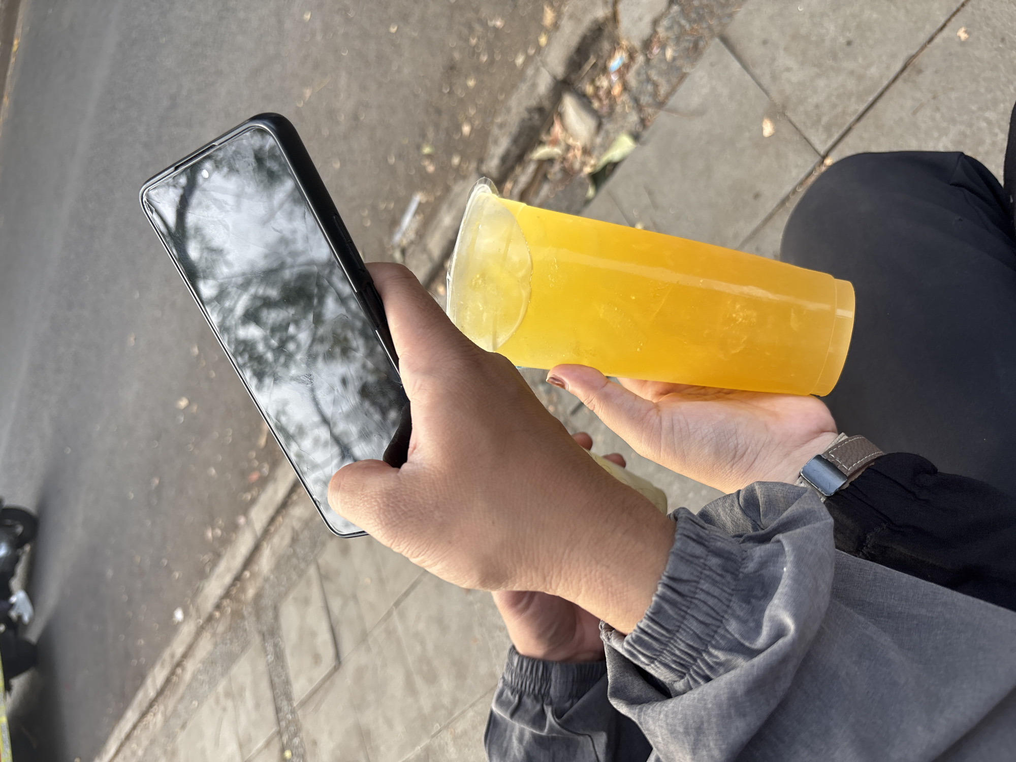 In Cirebon, Indonesia, a person is holding a smartphone and a plastic cup of yellow liquid. The person is wearing a grey jacket with elastic cuffs and black pants. The smartphone is in the person's left hand, and the cup is in the right. The background shows a sidewalk, a street, and a vehicle. The scene appears to be outdoors, likely on a street in Cirebon.