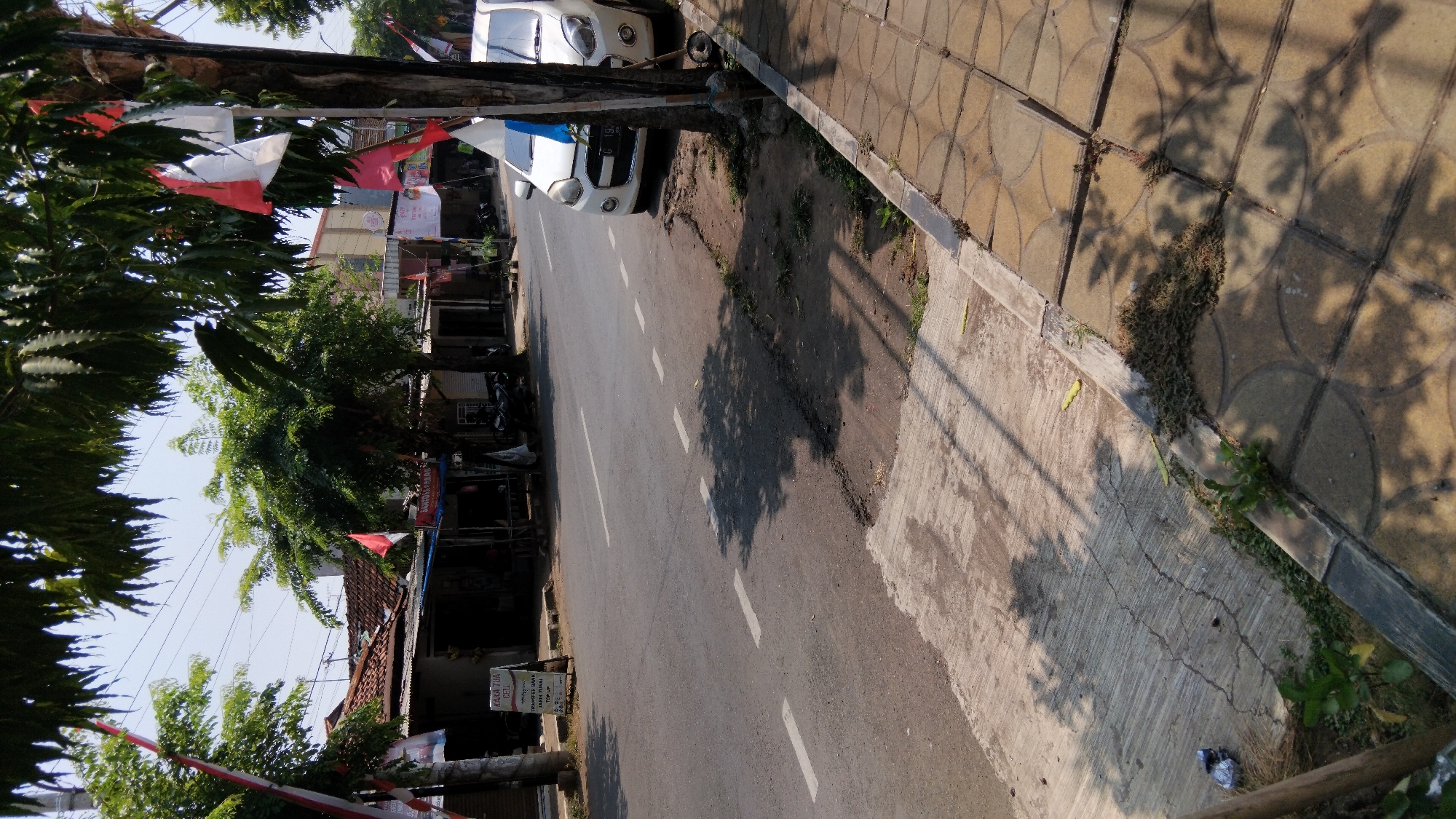 The image depicts a street scene in Proyonangg, Indonesia. A white car is parked on the right side of the road, close to a tree adorned with Indonesian flags. The road itself is asphalt with dashed white lines marking the lanes. To the left of the road, a sidewalk made of concrete and patterned tiles is visible, bordered by some overgrown vegetation. Across the street, there are several small buildings, likely shops, with awnings and some signage, including one that reads "SAKA TUA CELL". Motorcycles are parked in front of some of these establishments. The environment appears to be sunny, with shadows cast by trees on the road and sidewalk. The overall impression is a typical street in a residential or commercial area in Indonesia.