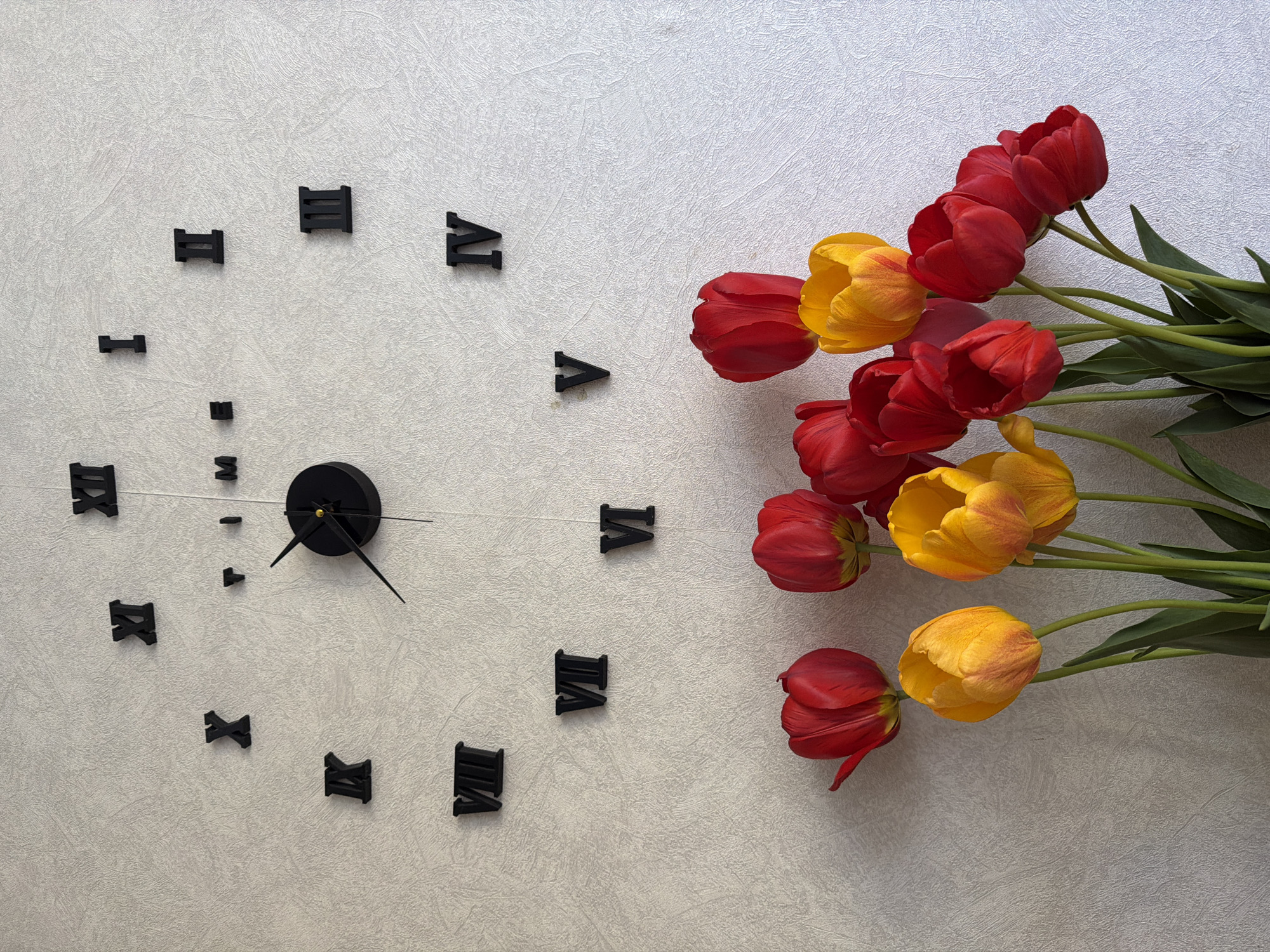 A top-down shot shows a white wall, on which is affixed a DIY-style clock made of black, individually applied Roman numerals and a central clock mechanism with two hands. The clock face is incomplete, lacking several numeral components. To the right of the clock, a bouquet of red and yellow tulips is positioned, with their stems and leaves extending towards the upper portion of the frame.