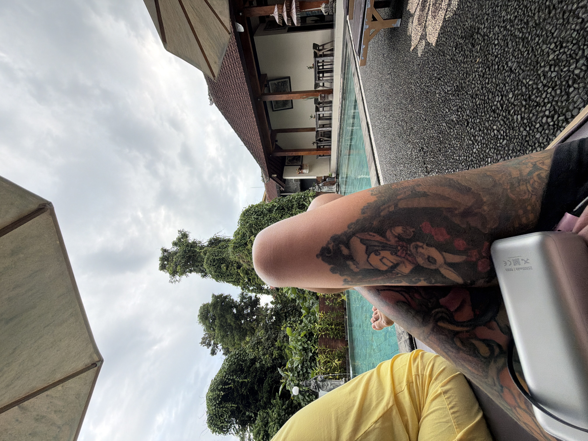 In a tranquil outdoor setting, a person relaxes poolside in the district of Ubud, Indonesia. The person, whose lower body is prominently featured, is adorned with extensive tattoos on their legs. They are wearing bright yellow pants. A silver portable charger rests on their lap.
The scene is set around a bright blue swimming pool, bordered by a textured grey stone path. Lush green foliage and tall trees create a vibrant backdrop, suggesting a tropical environment. In the distance, a building with a reddish-brown tiled roof, supported by wooden beams, houses tables and chairs, likely an outdoor dining or lounge area. The sky above is overcast, with diffused light suggesting a cloudy day. A portion of a large light-colored umbrella is visible in the upper left corner of the frame, providing shade.