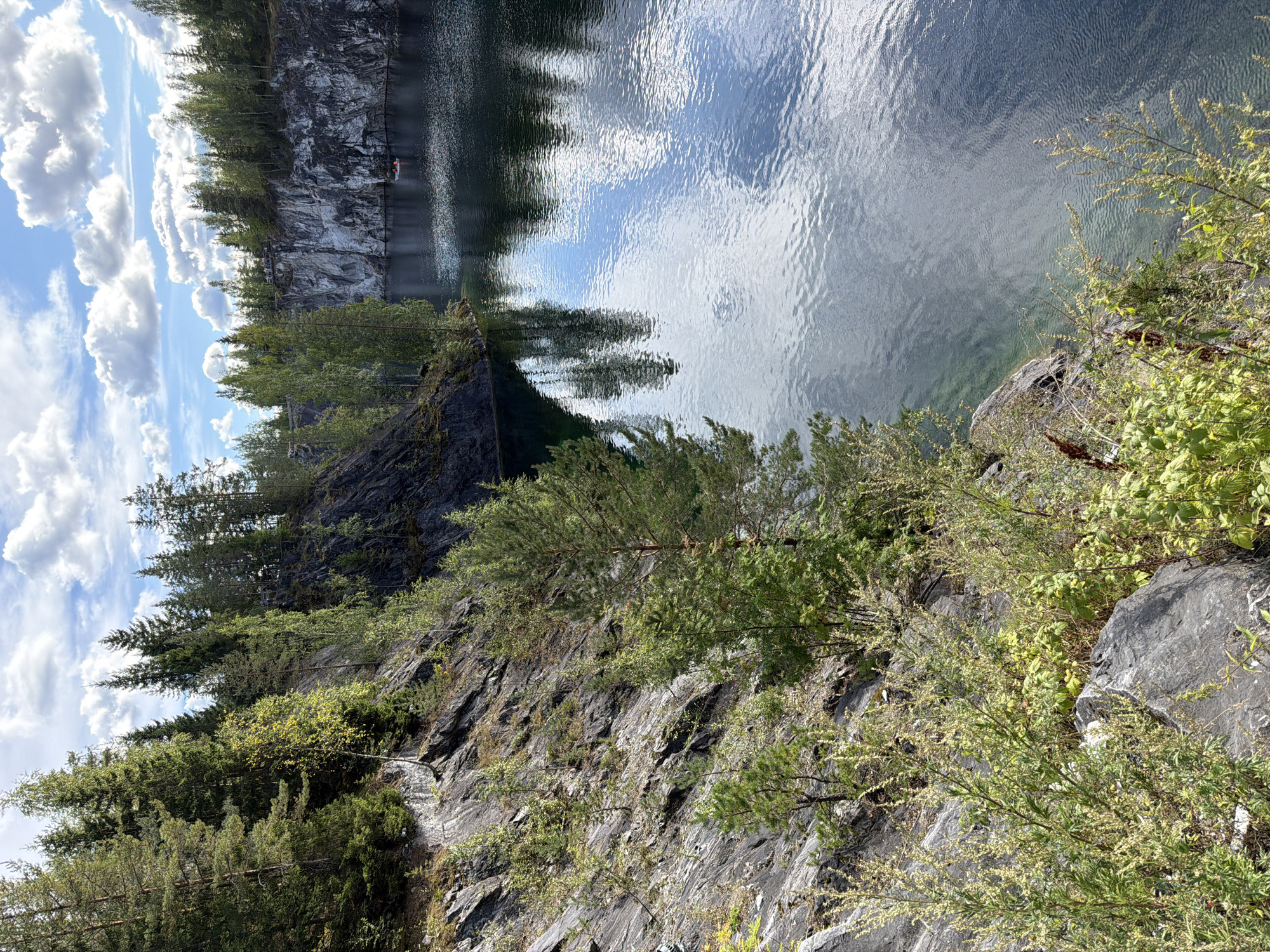A wide-angle, natural light shot captures a scenic view of a tranquil lake nestled between steep, rocky cliffs covered in dense evergreen trees. The water, dark and reflective, mirrors the cloudy blue sky and the surrounding greenery. In the distance, on the lake, a small white boat with two individuals can be observed. The foreground features a rocky embankment overgrown with lush green vegetation, including small trees and shrubs. The overall impression is one of natural beauty and serenity in an outdoor environment.