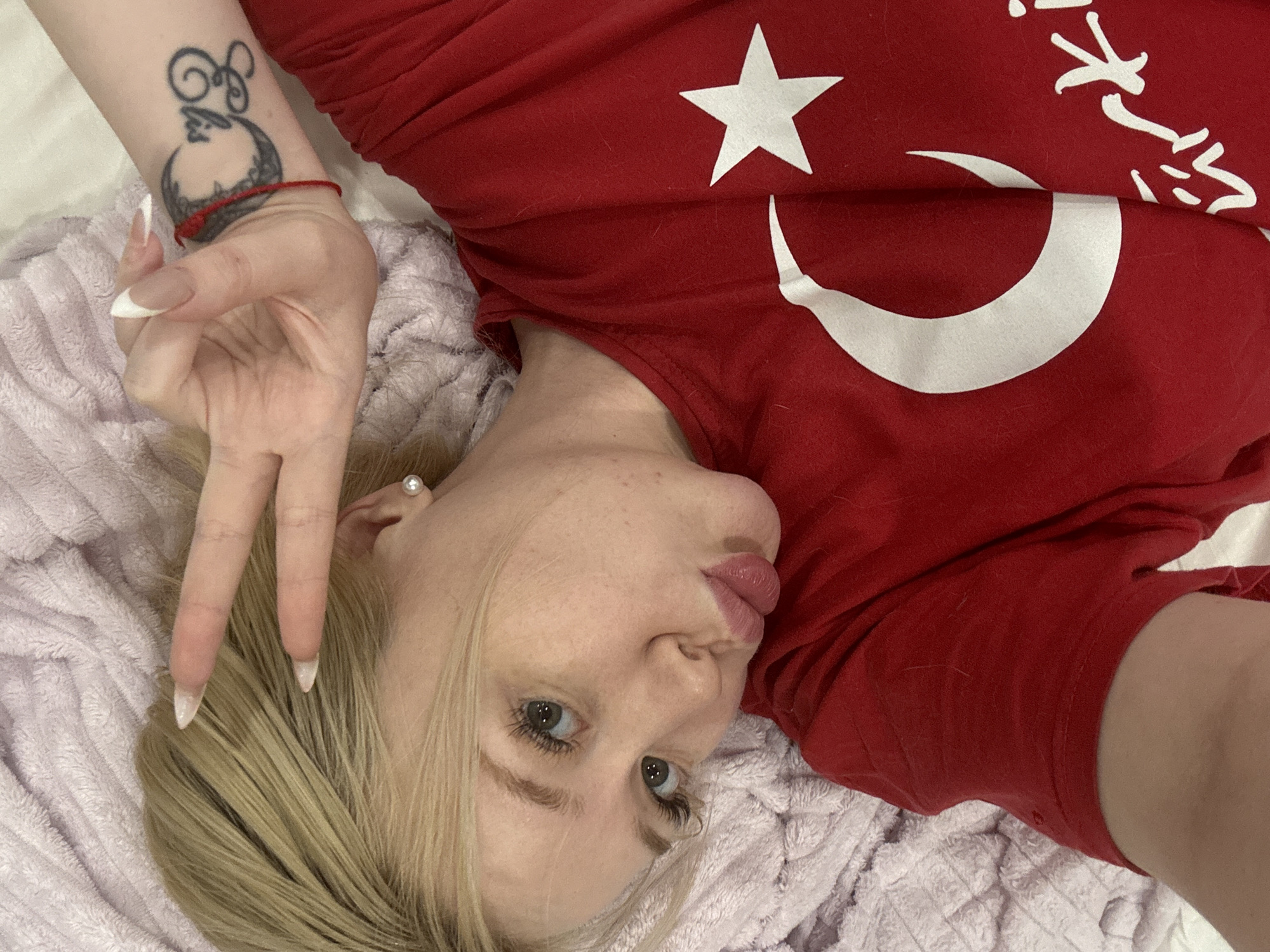 An overhead shot shows a blonde woman lying down on a light purple blanket. She is wearing a red t-shirt with a white crescent moon and star, the symbol of the Turkish flag. Her right hand is raised, forming a peace sign with her fingers. She has a tattoo on her left forearm with a crescent moon and what appears to be Arabic script. The woman is looking at the camera with her lips pursed. The overall setting appears to be indoors, likely a bedroom.