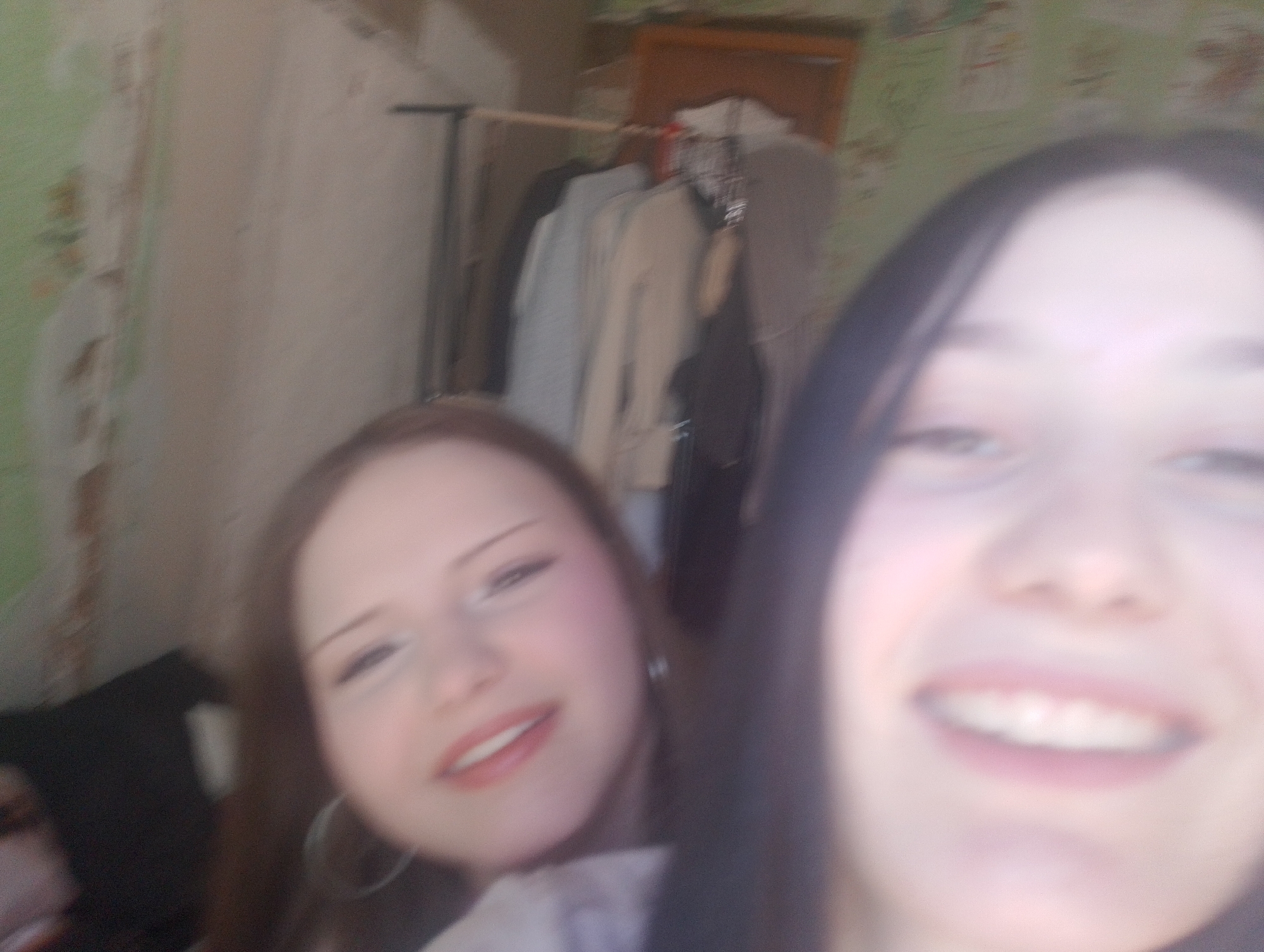 The image shows two young women in an indoor setting. The woman on the left has brown hair and is wearing hoop earrings. She is smiling. The woman on the right has dark hair and is also smiling, but her face is closer to the camera and slightly out of focus.
In the background, there is a clothing rack with several garments hanging on it. A wooden cabinet or shelf is visible above the clothing rack. The wallpaper appears to have a faded floral pattern.
Based on the image alone, there is no evidence to suggest that the location is Фоки, Russia.